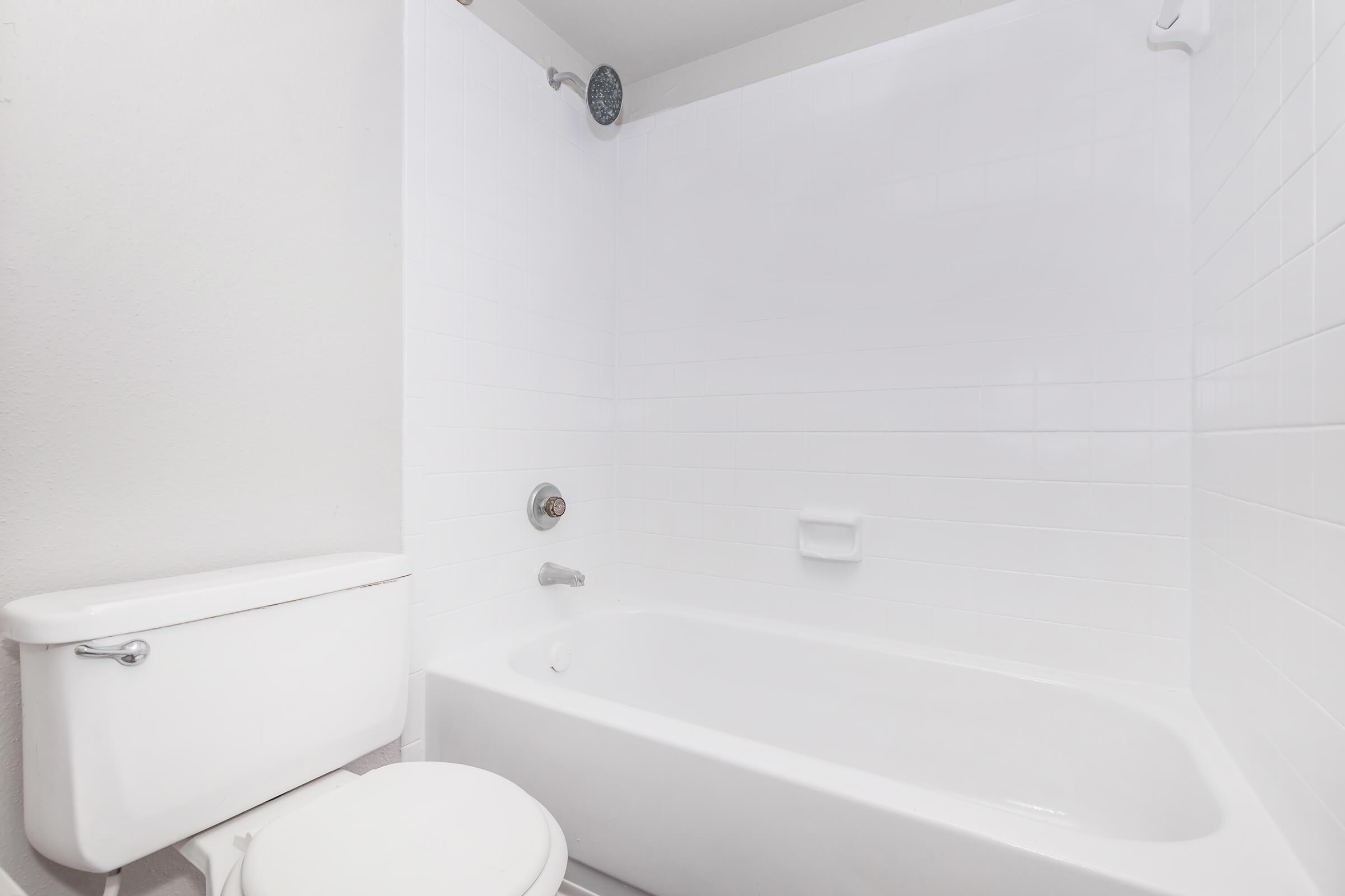 A clean, modern bathroom featuring a white bathtub with a showerhead, a white toilet, and tiled walls. The space has a minimalistic design with bright lighting, emphasizing a fresh and uncluttered appearance.