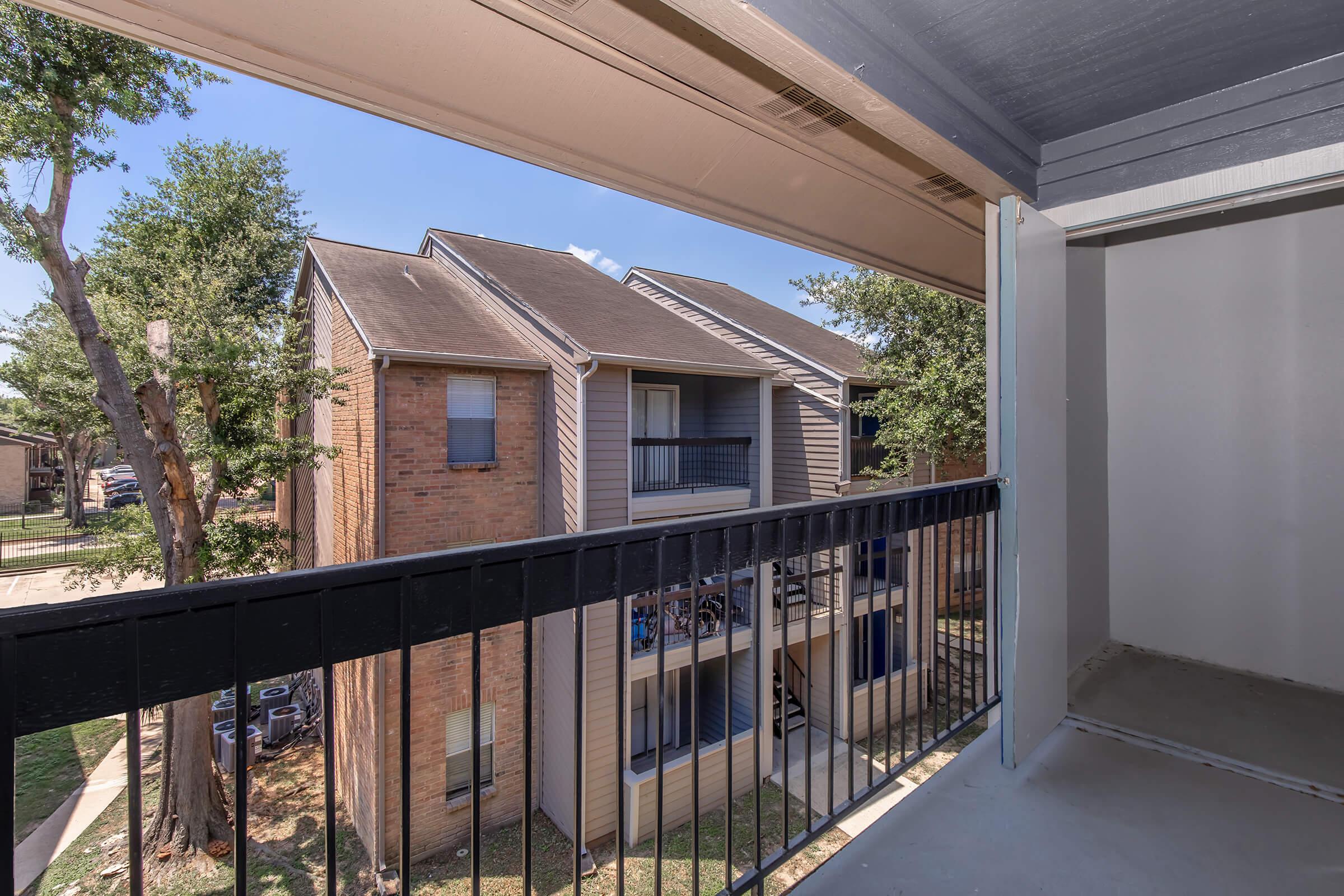 View from a balcony of an apartment building, showcasing a neighboring structure with brick exterior and balconies. A tree is visible nearby, and the sky is clear. The balcony features a railing and an empty adjacent storage space.