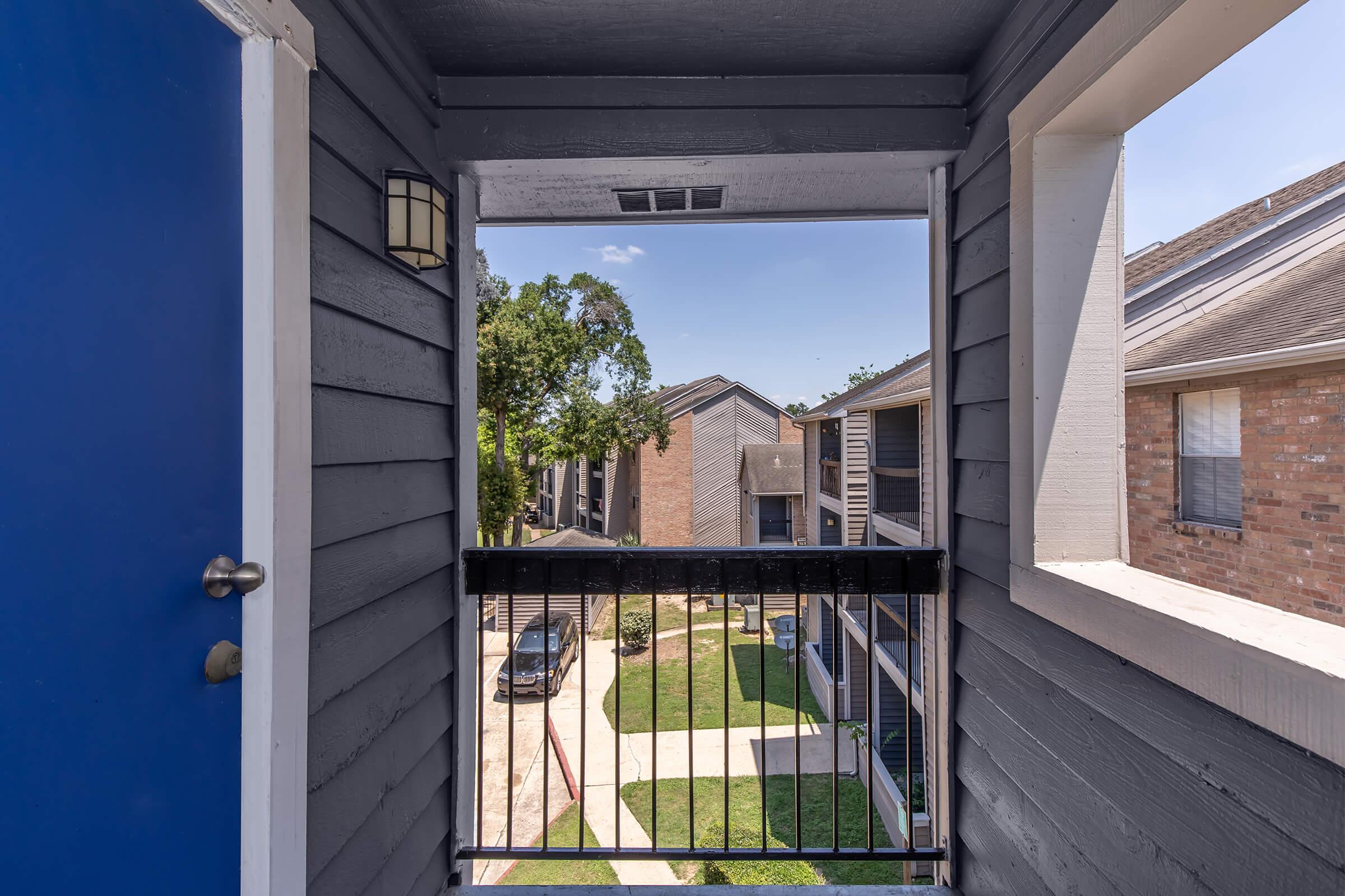 A view from a second-floor balcony featuring a blue door on the left, with a railing framing the scene. Below, a tree and landscaped walkway are visible, leading to other apartment buildings in the background under a clear blue sky.