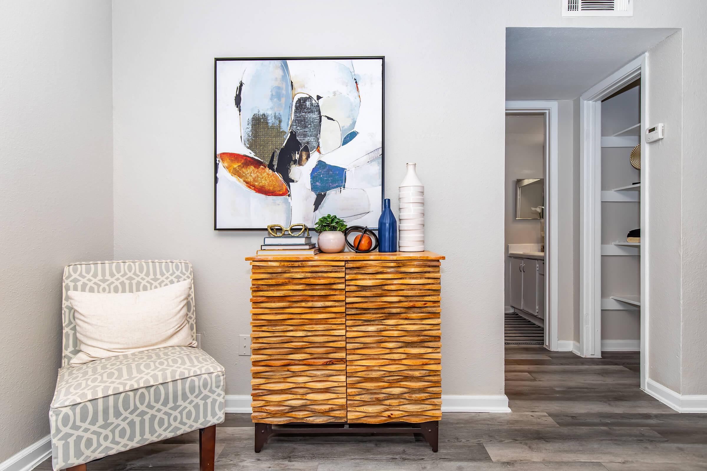 A cozy corner featuring a textured wooden cabinet with decorative items on top, a potted plant, and colorful accents. Next to it, a patterned upholstered chair provides seating. In the background, a doorway leads to a hallway with shelves, and a piece of abstract art hangs on the wall above the cabinet.