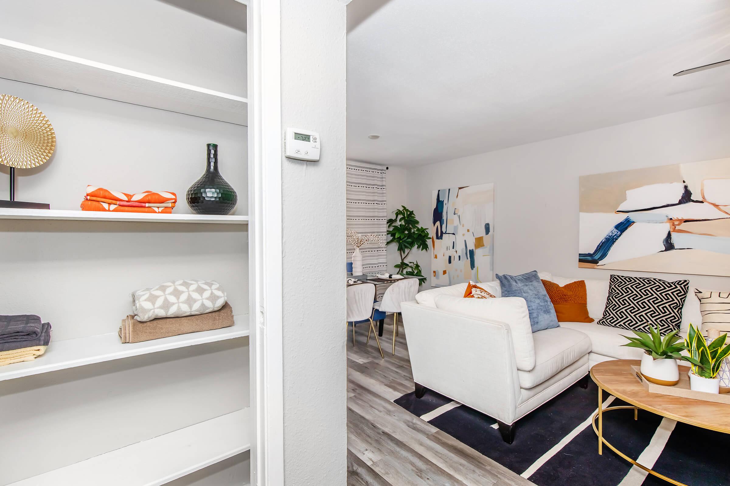A cozy living room with a white couch adorned with decorative pillows, a round coffee table, and a stylish rug. Shelves on the left display neatly folded towels and decorative items. A dining area in the background features a plant and an abstract artwork on the wall, creating a bright and inviting atmosphere.