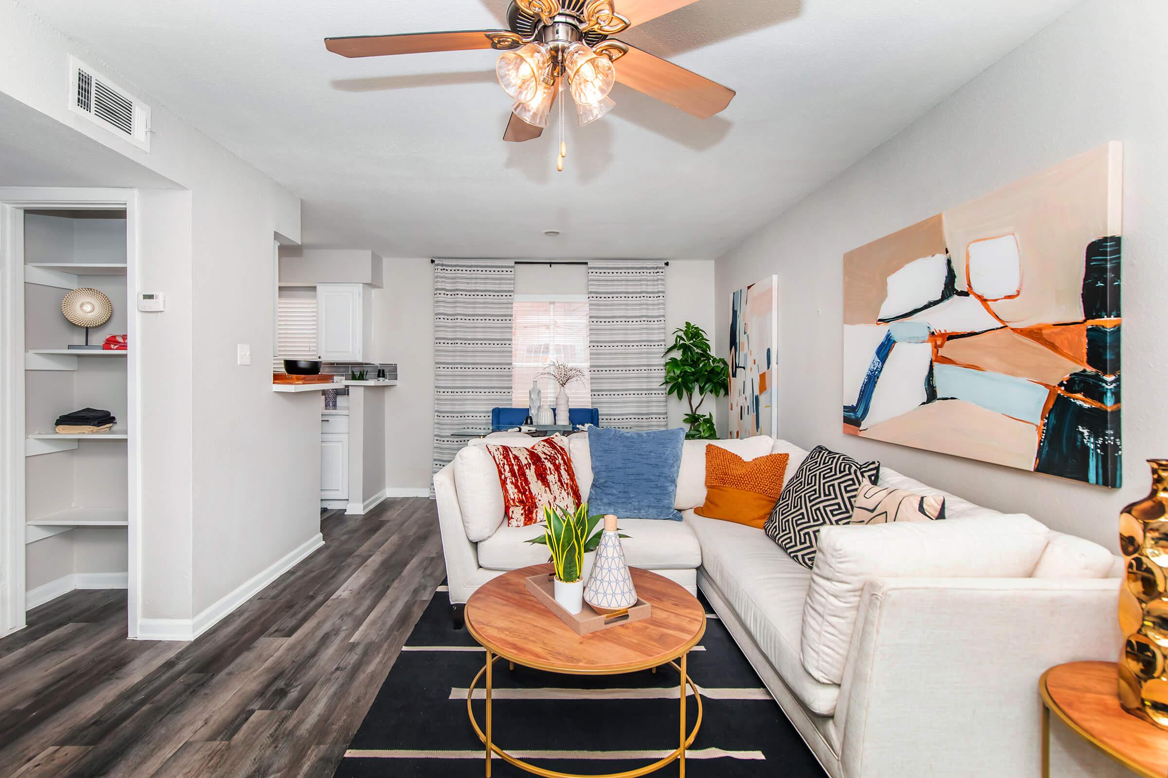 A modern living room featuring a white couch with colorful throw pillows, a round wooden coffee table, and a decorative plant. The space has a ceiling fan and a large piece of artwork on the wall. There are shelves on the left and a view into a kitchen area visible in the background.