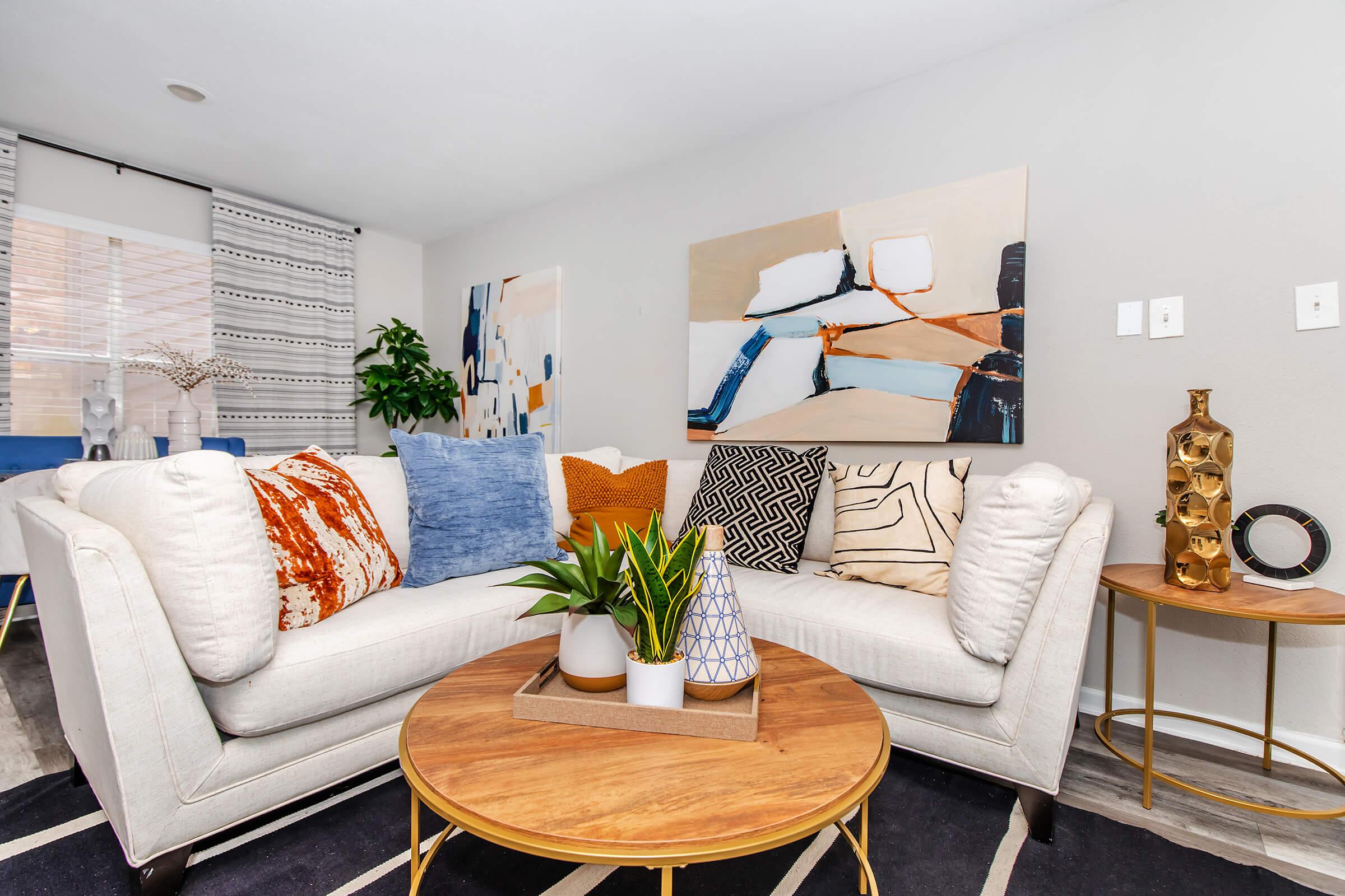 A cozy living room featuring a white sectional sofa adorned with colorful pillows in blue, orange, and patterned designs. A round wooden coffee table displays a small plant and decorative vases. The walls are decorated with modern art pieces, and there’s a plant in the corner, adding a touch of greenery to the space.