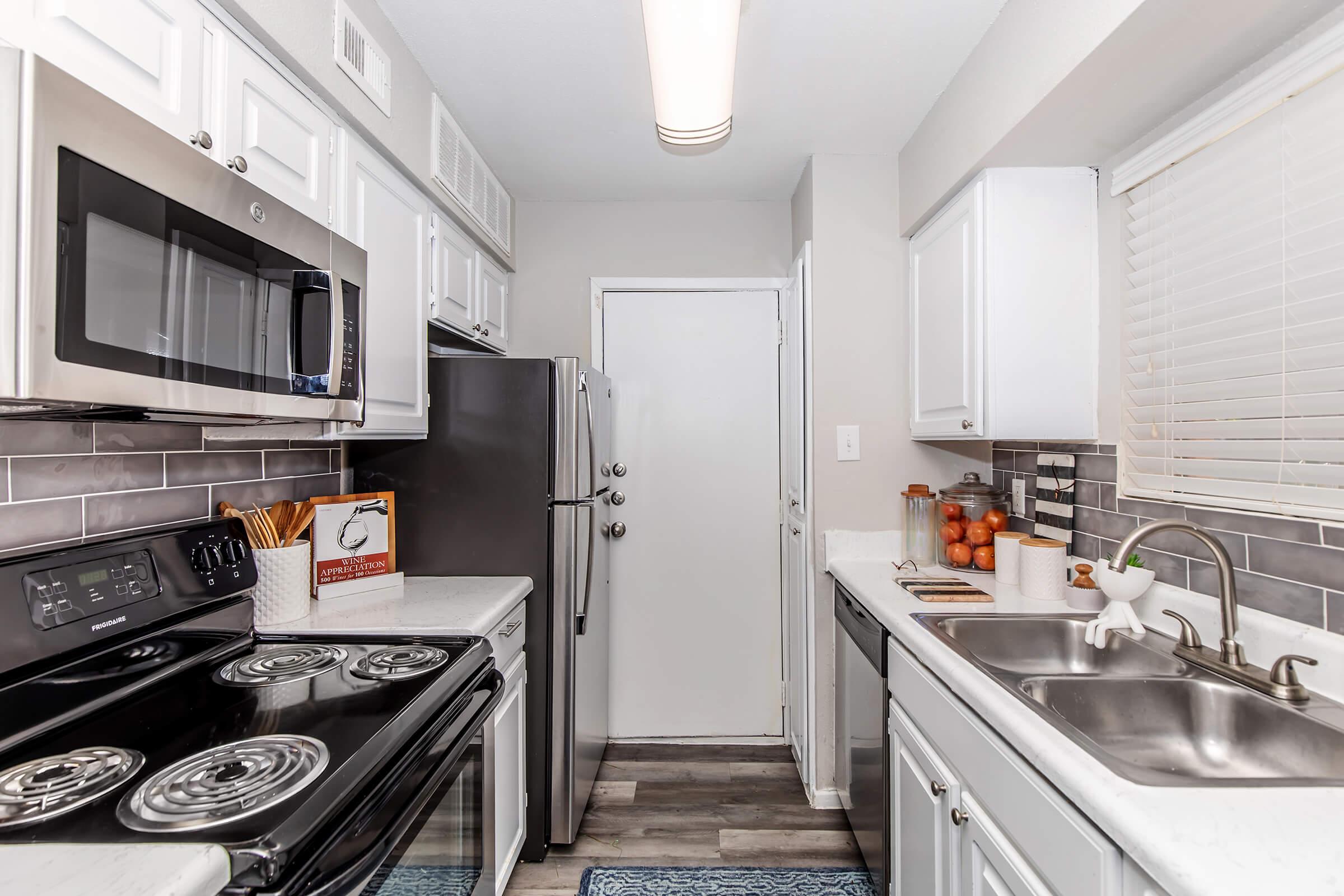 A modern kitchen featuring stainless steel appliances, including a microwave, refrigerator, and stove. The space has white cabinets, a double sink, and a window with blinds. A bowl of oranges sits on the countertop, and a decorative mat adds a cozy touch to the floor.