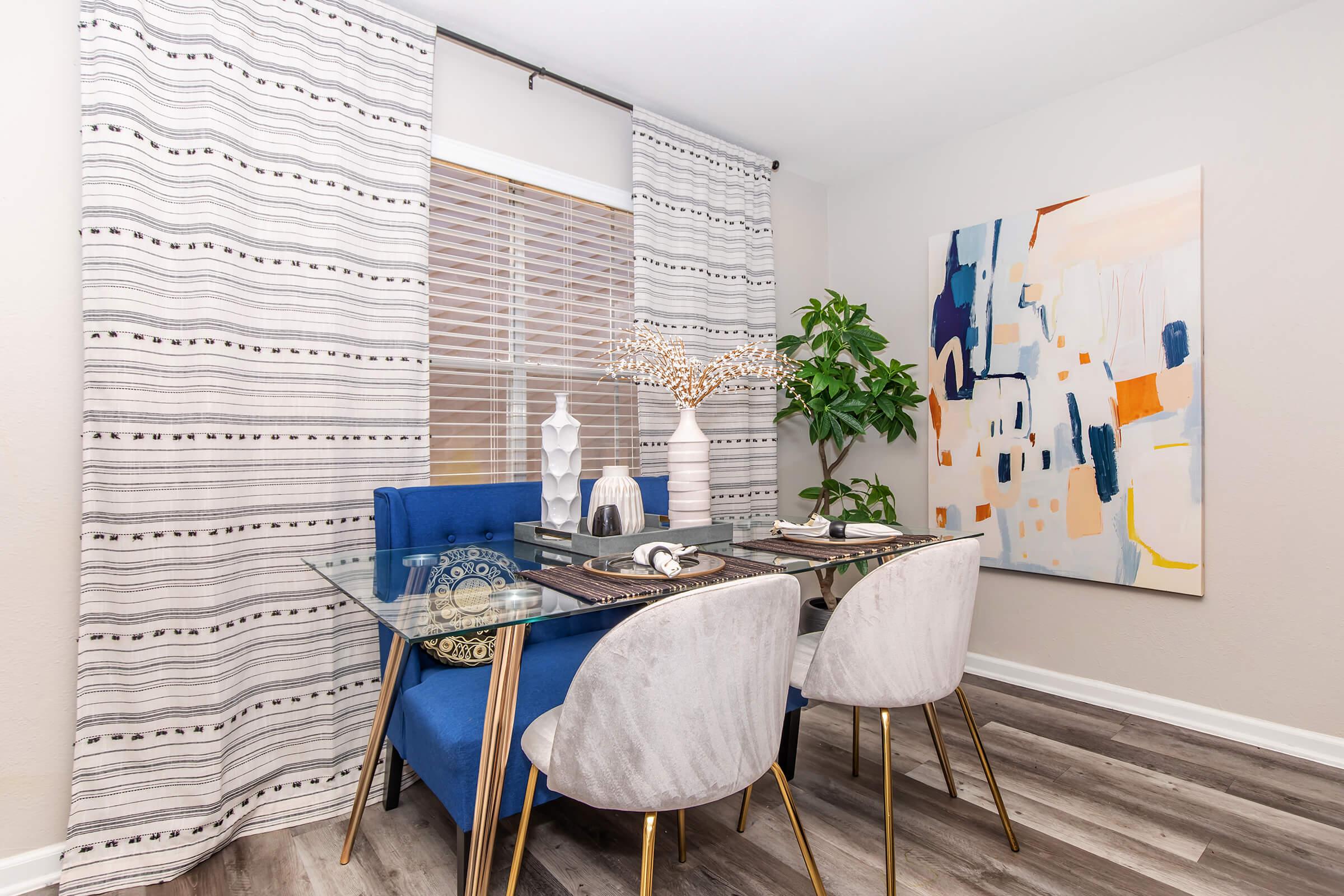 A modern dining area featuring a glass table with two stylish gray chairs and a blue upholstered bench. The space is decorated with a colorful abstract painting, a potted plant, decorative vases, and drapes with a subtle pattern, all against light-colored walls and hardwood flooring.