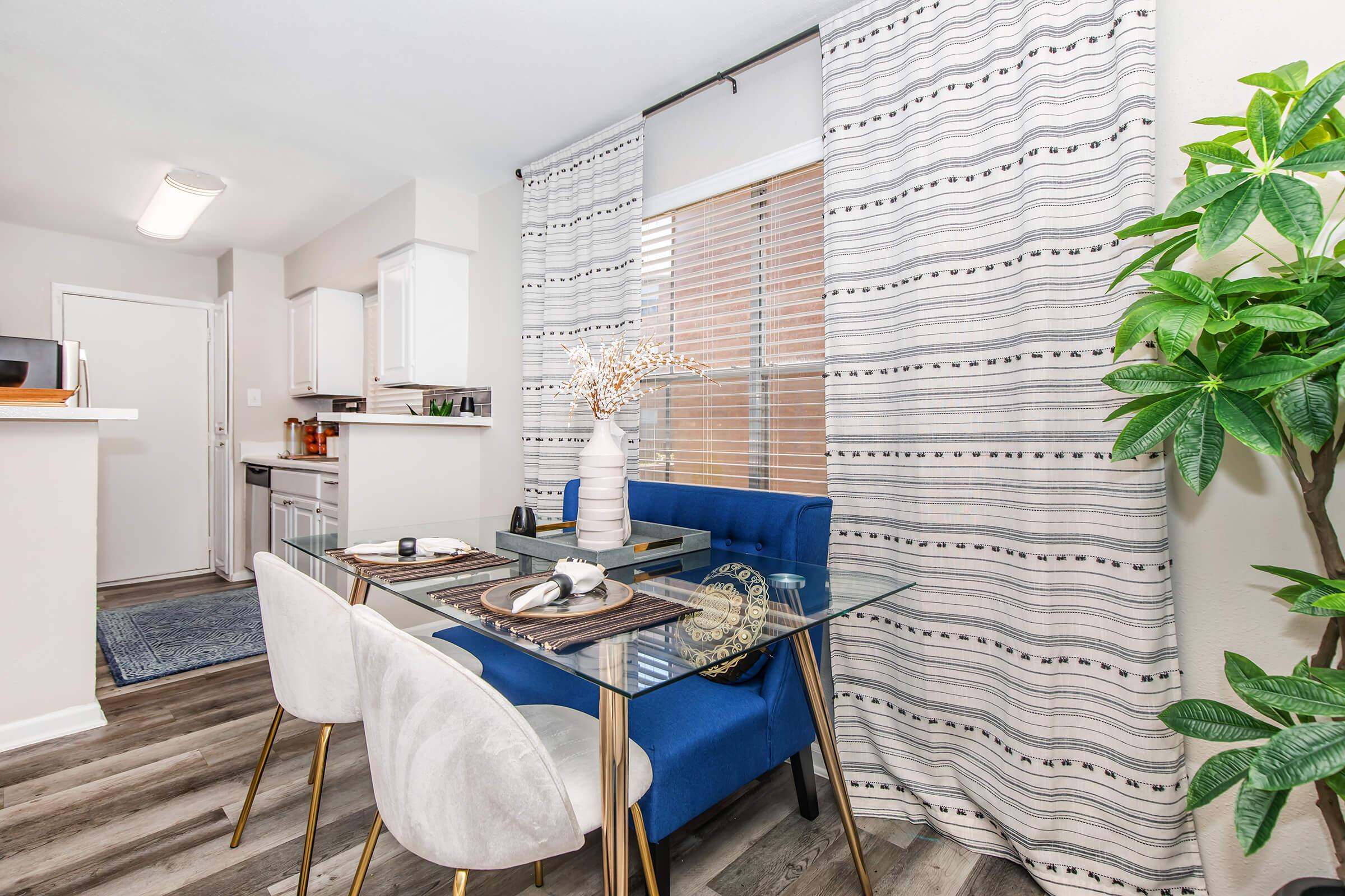 A modern dining area featuring a glass table with place settings, a blue upholstered dining chair, and stylish white chairs. Natural light fills the space through curtains. A plant adds a touch of greenery, and the open kitchen area is visible in the background, enhancing a cozy, inviting atmosphere.