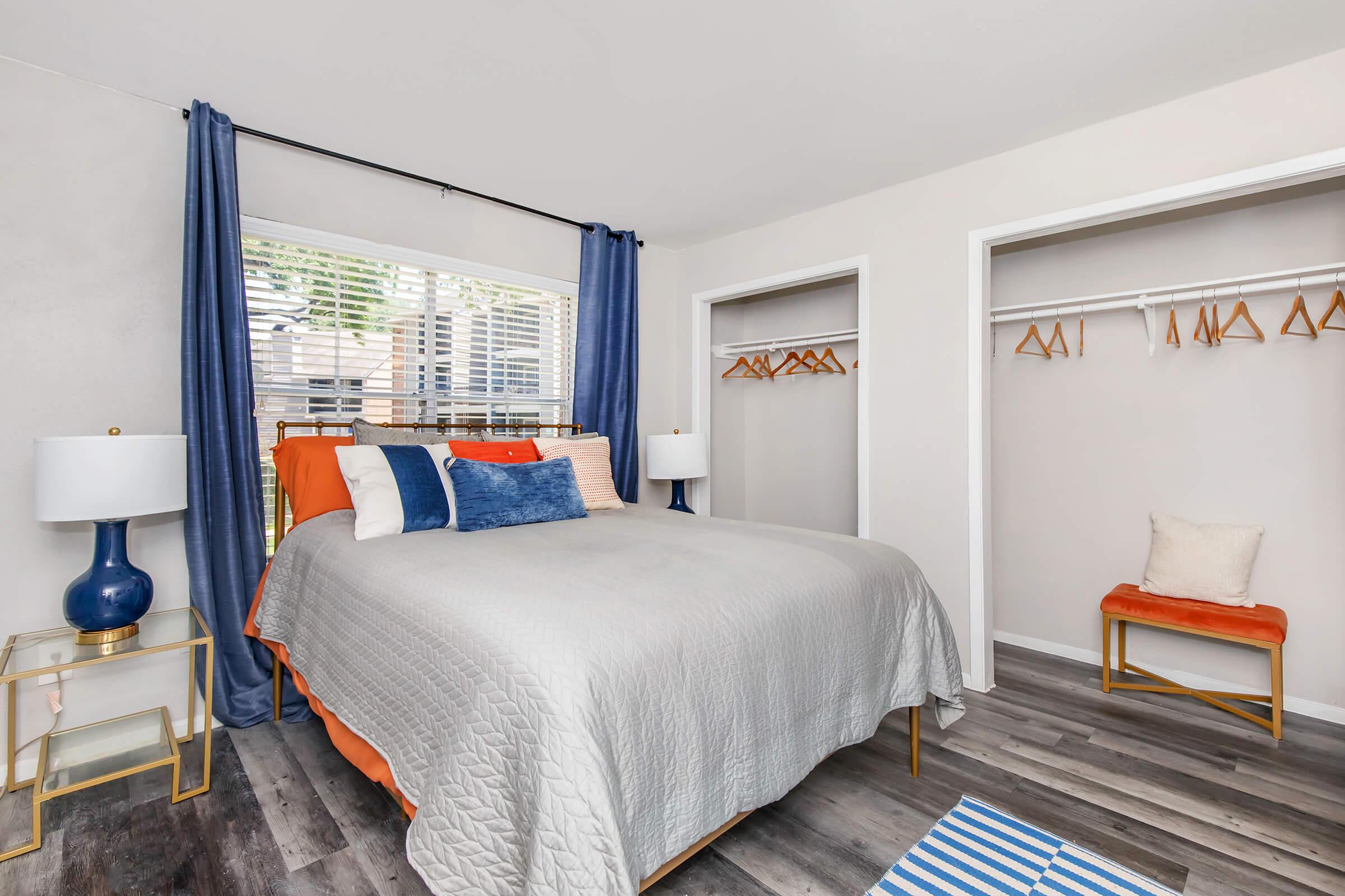 A cozy bedroom featuring a queen-sized bed with gray bedding and colorful accent pillows. One side has a nightstand with a blue lamp, while two closets with hanging space are visible. Natural light comes through window curtains, creating a warm atmosphere. A small orange chair adds a pop of color to the room.