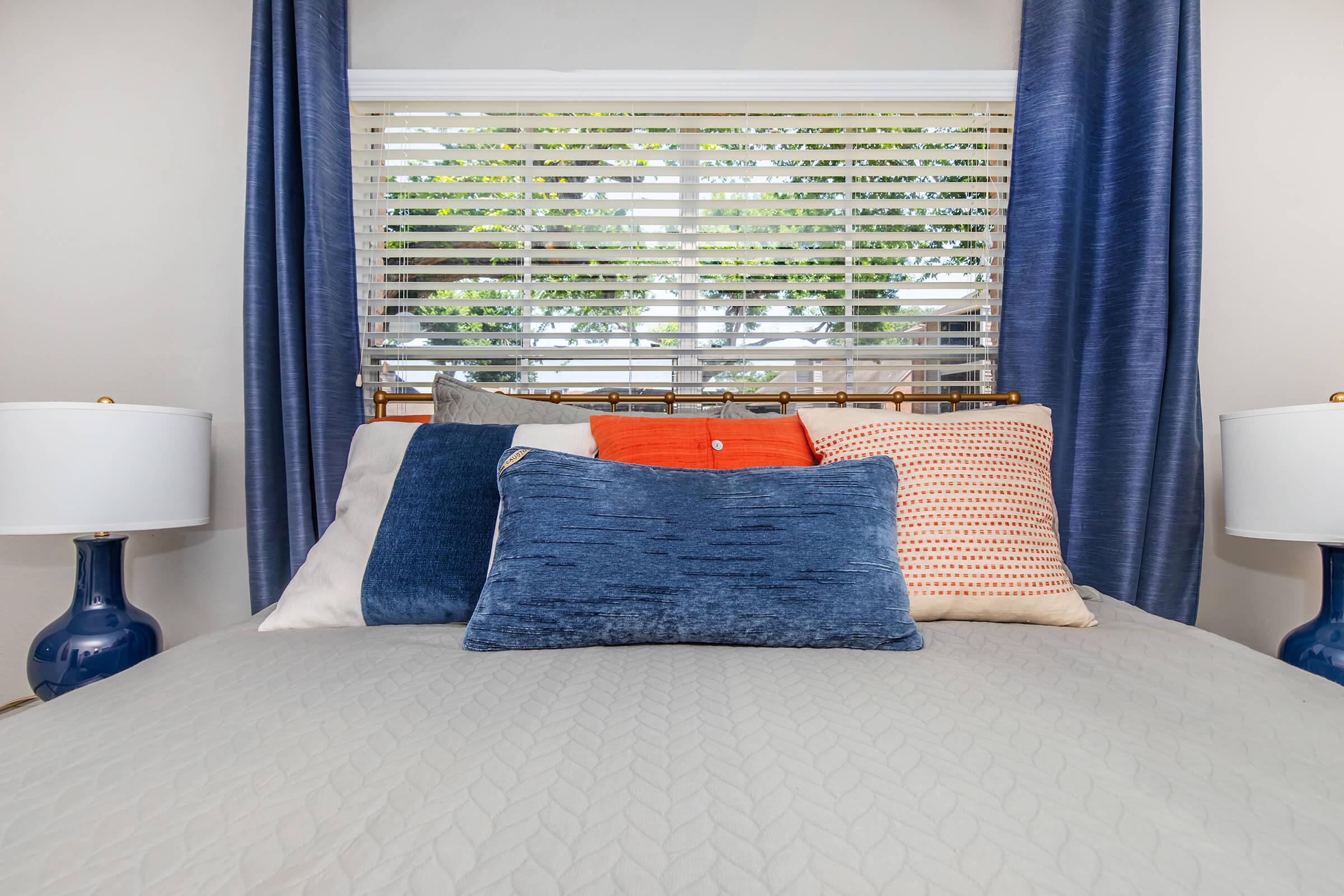 A neatly made bed featuring decorative pillows in various colors, including blue, orange, and white. There are two bedside lamps with blue bases on either side of the bed. Behind the bed, a window with blinds allows natural light to filter in, showcasing some greenery outside.