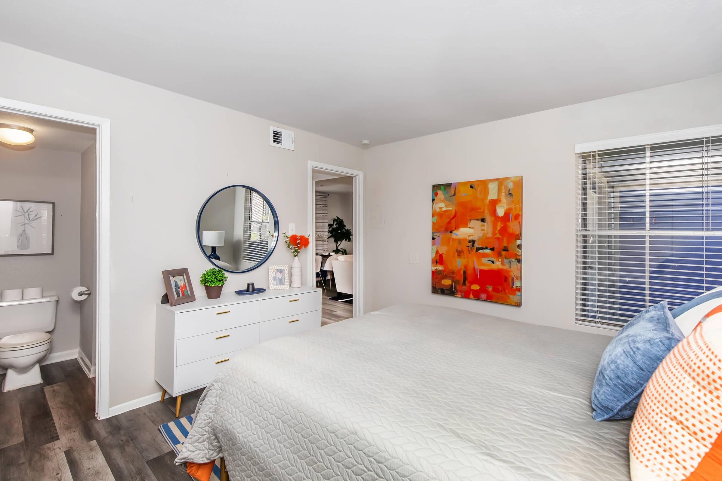 A well-lit bedroom featuring a gray bedspread, decorative pillows, and an orange abstract painting on the wall. A round mirror is above a dresser with framed photos and a plant. To the side, there's a door leading to a bathroom. Large windows provide natural light, enhancing the cozy atmosphere.