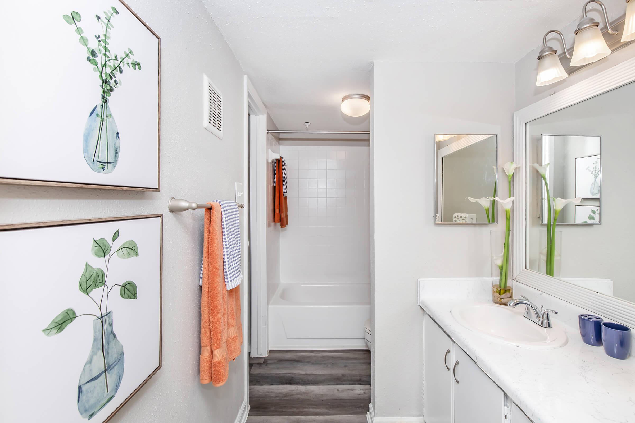 A modern bathroom featuring light gray walls, a white vanity with a sink, and a large mirror. Two framed botanical prints adorn the wall. An orange towel hangs on a rack, and there are decorative plants on the vanity. The shower area is partially visible in the background.
