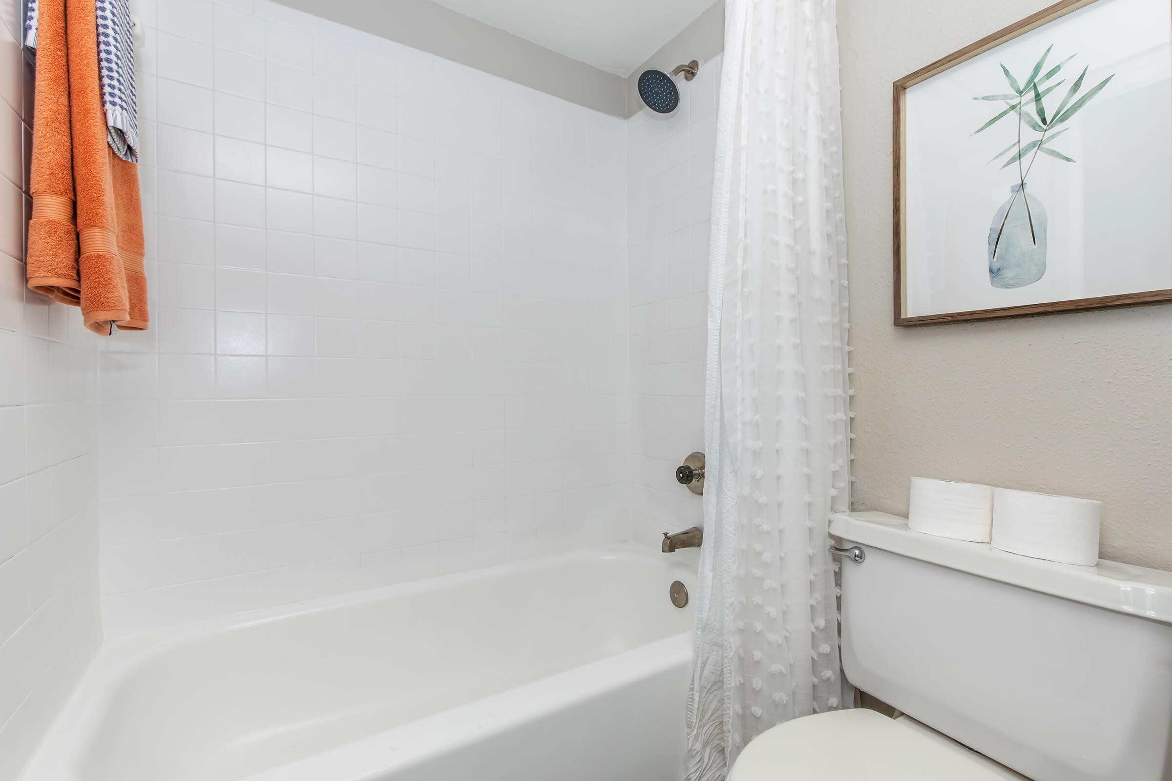 A clean, modern bathroom featuring a white bathtub with a shower curtain, a shower head, and neatly arranged towels hanging nearby. A decorative wall art piece showing a plant in a vase is mounted on the wall, alongside a toilet with two toilet paper rolls placed on top.