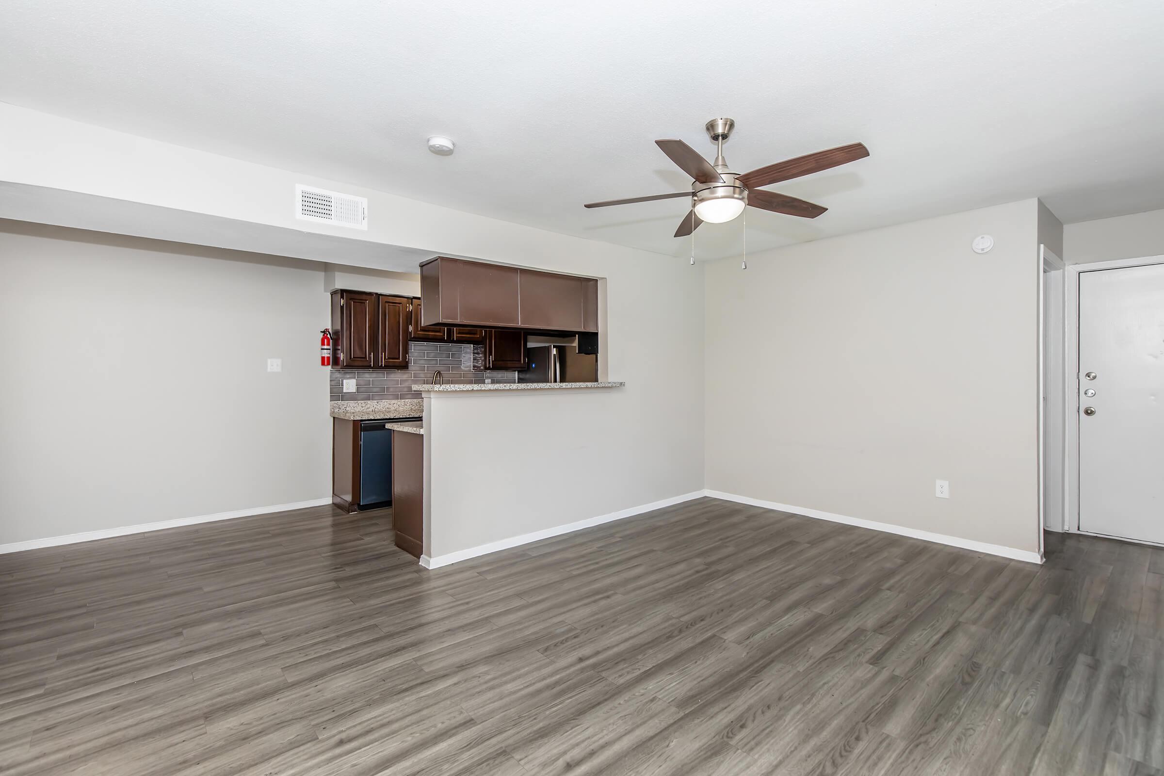 Spacious living area with laminate flooring, featuring a ceiling fan and an open layout. The kitchen, partially visible, has dark cabinetry and a countertop that extends to the living space. Neutral-colored walls enhance the bright, airy atmosphere. A doorway leads to the entrance of the room.