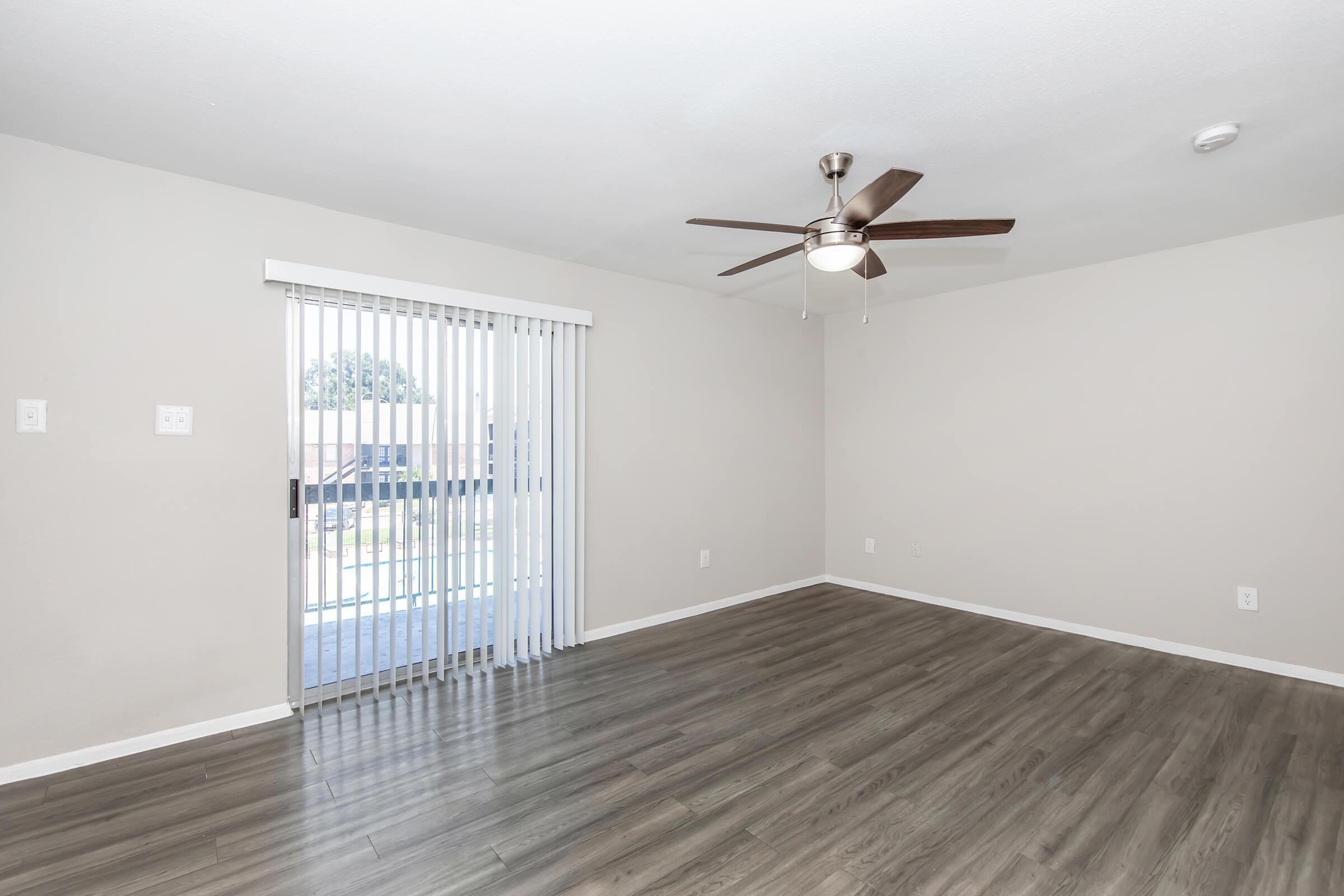 A spacious, empty living room featuring light-colored walls and a ceiling fan. Large sliding glass doors with white vertical blinds allow natural light, and the floor is covered in dark laminate wood. There are no furnishings, creating an open and airy atmosphere.
