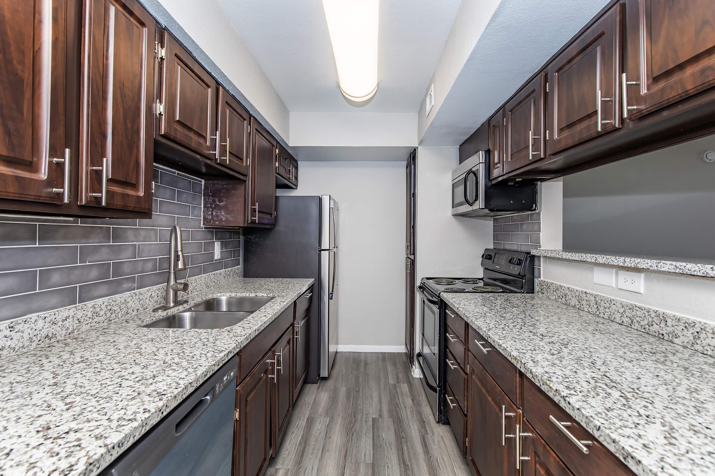 Modern kitchen featuring dark wooden cabinets, granite countertops, and stainless steel appliances. The layout includes a refrigerator, microwave, and oven, with a sleek tile backsplash and ample countertop space. Natural lighting enhances the contemporary design.