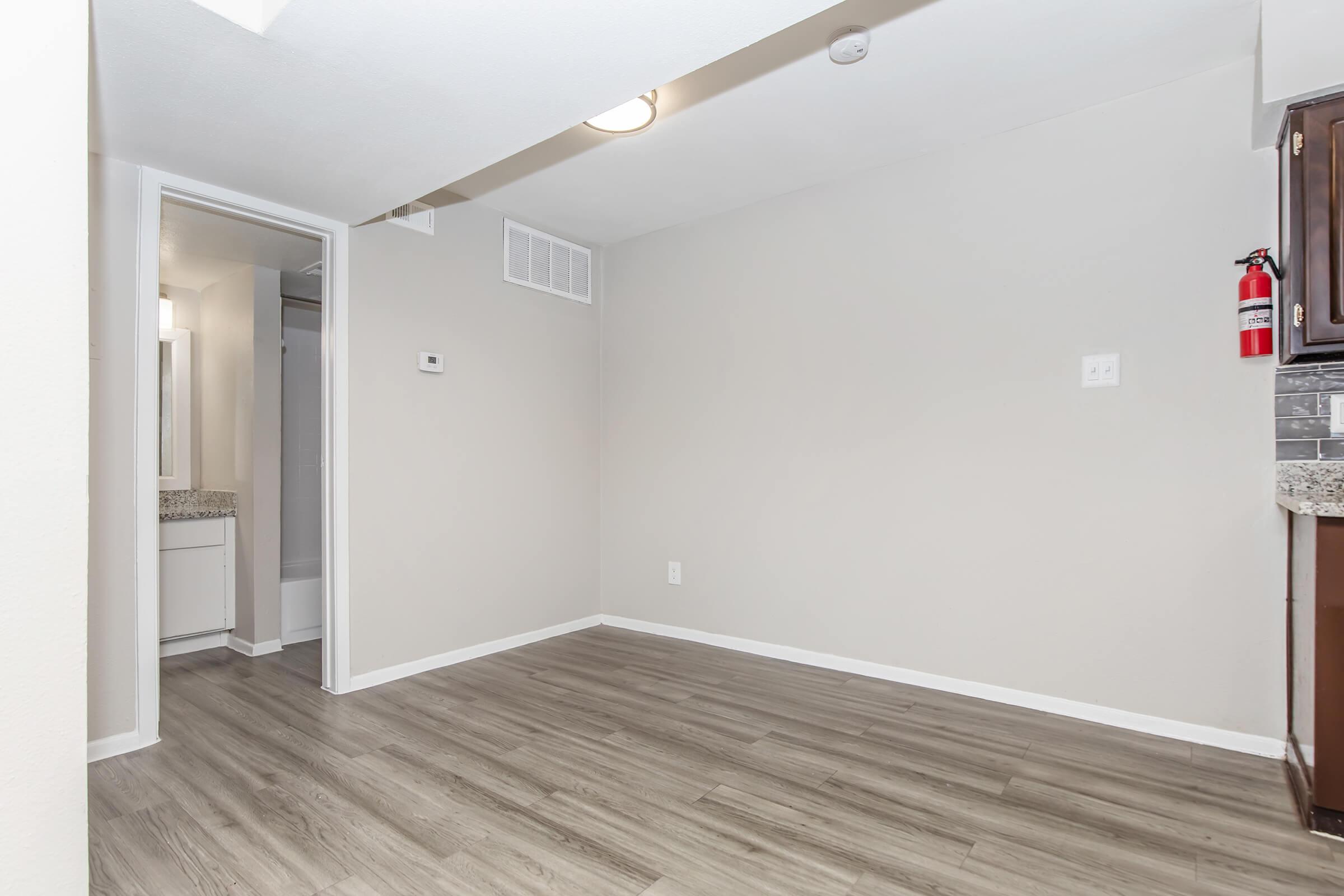 A clean and bright room featuring light-colored walls and a wood-look floor. There is an open doorway leading to a bathroom area, with a small wall-mounted thermostat visible. The kitchen area shows part of a textured countertop and dark cabinetry. A fire extinguisher is mounted on the wall.