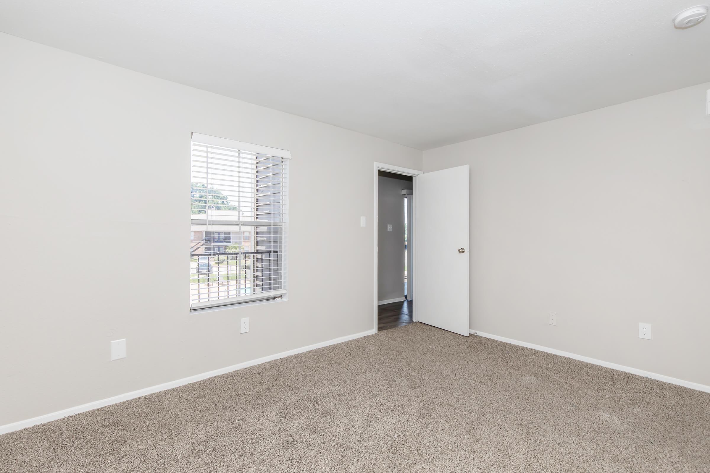 A clean, empty room with light beige walls and carpet. A window with white blinds lets in natural light, and there is a white door leading to another area. The room is unfurnished and appears spacious, with a minimalist aesthetic.