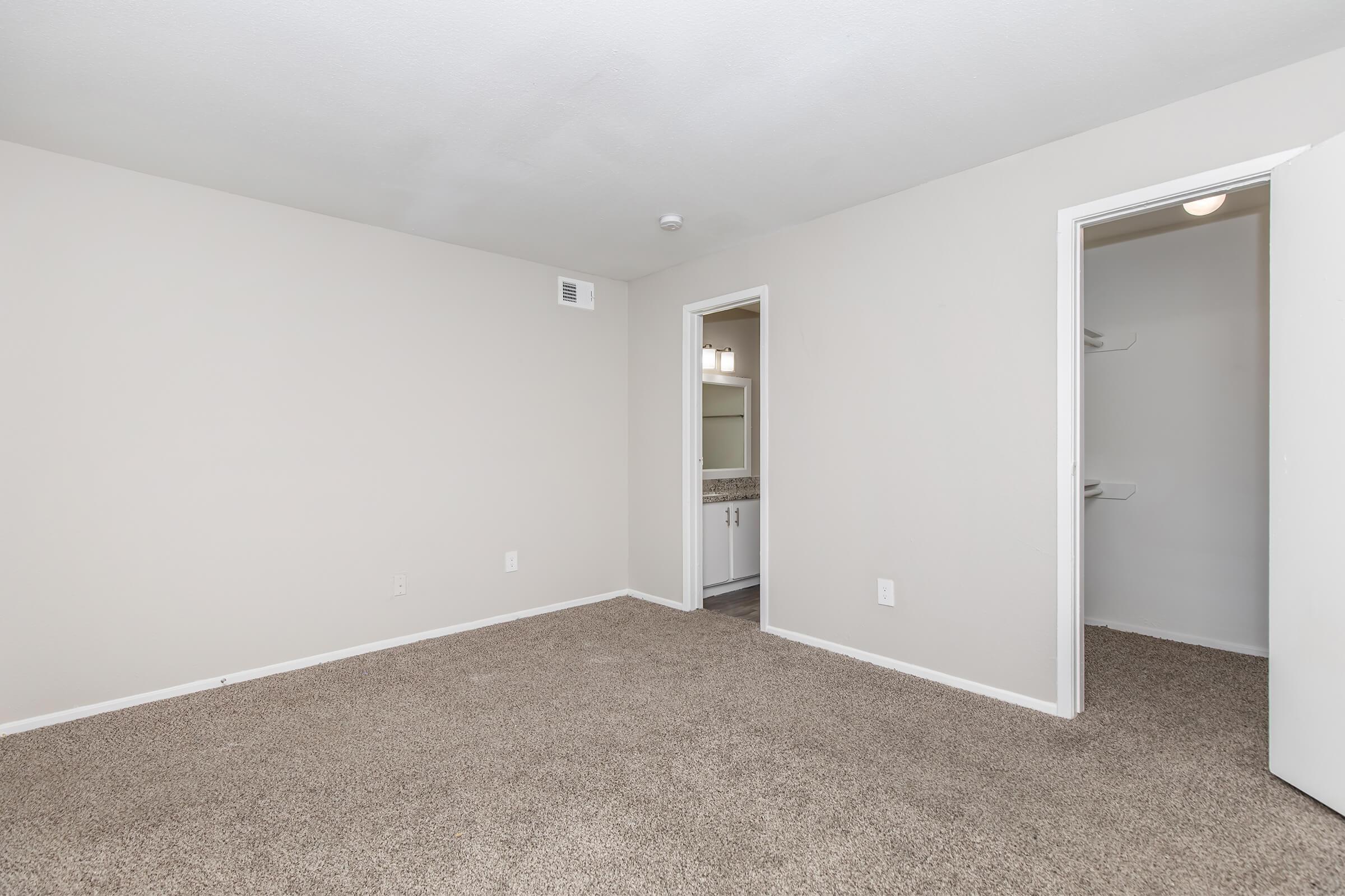 A spacious, empty room with beige walls and carpeted flooring. There is a doorway leading to a bathroom and another door on the right. The space is well-lit, with no furniture or decorations, showcasing a clean and neutral environment suitable for personal decoration.