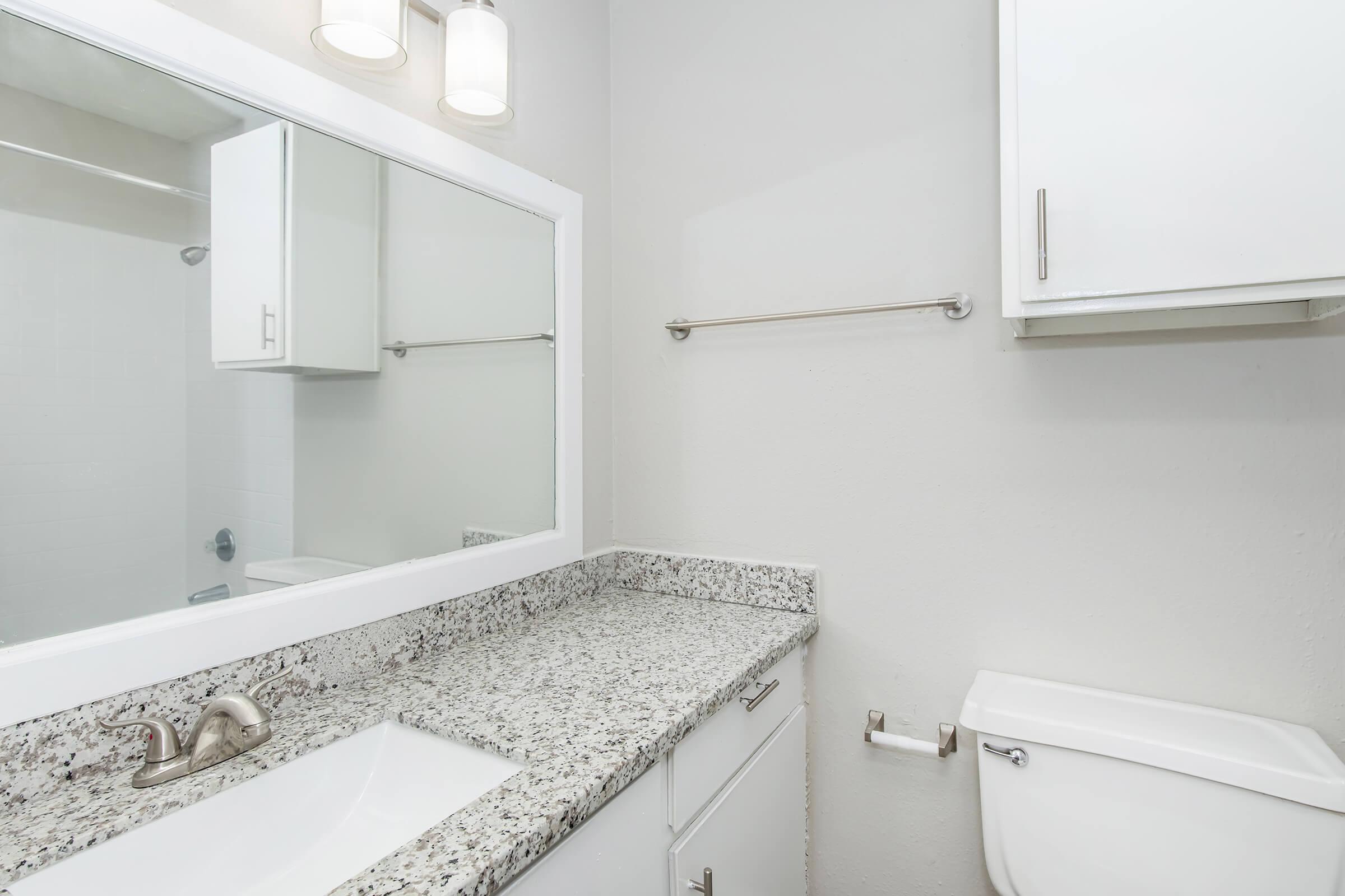 A clean and modern bathroom featuring a large mirror above a granite countertop with a sink, a wall-mounted towel bar, and a white toilet. The walls are painted a light color, and there are two light fixtures above the mirror, providing bright illumination.