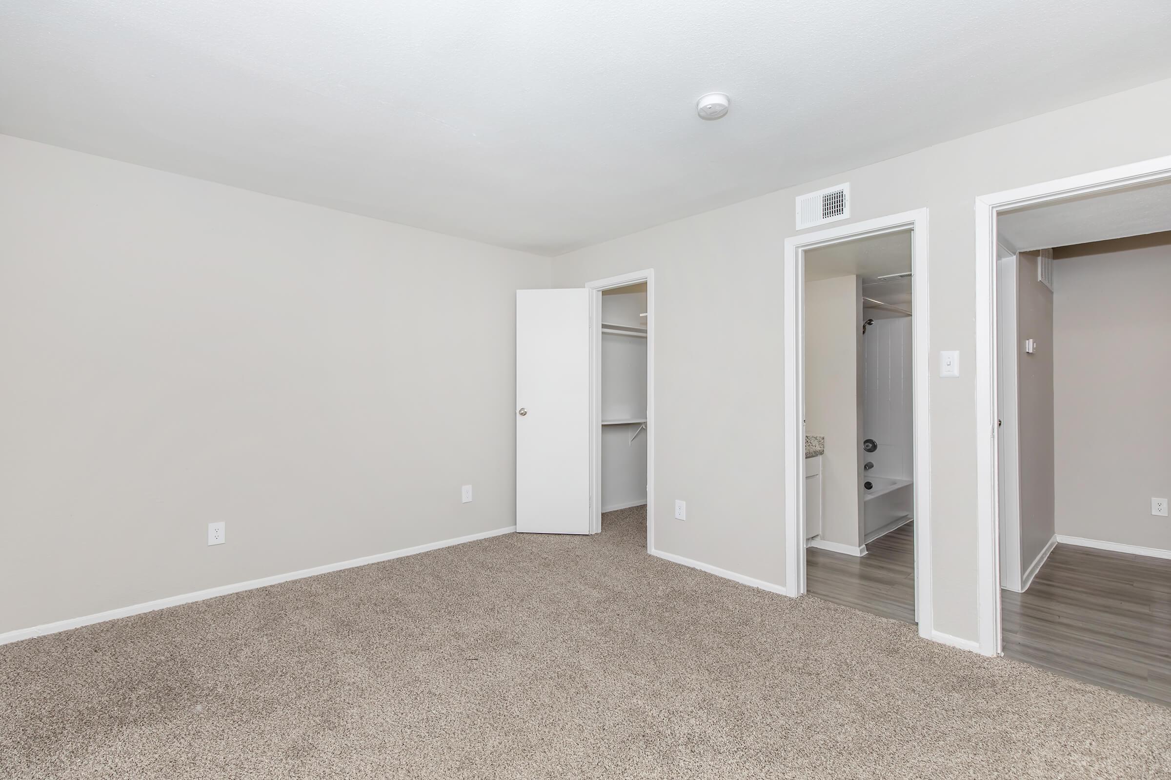 A spacious, empty room with light gray walls and tan carpet. There are two doorways leading to adjacent rooms, one with a closet space and another with an entrance to a bathroom. The area is well-lit with natural light, creating a bright and airy atmosphere.