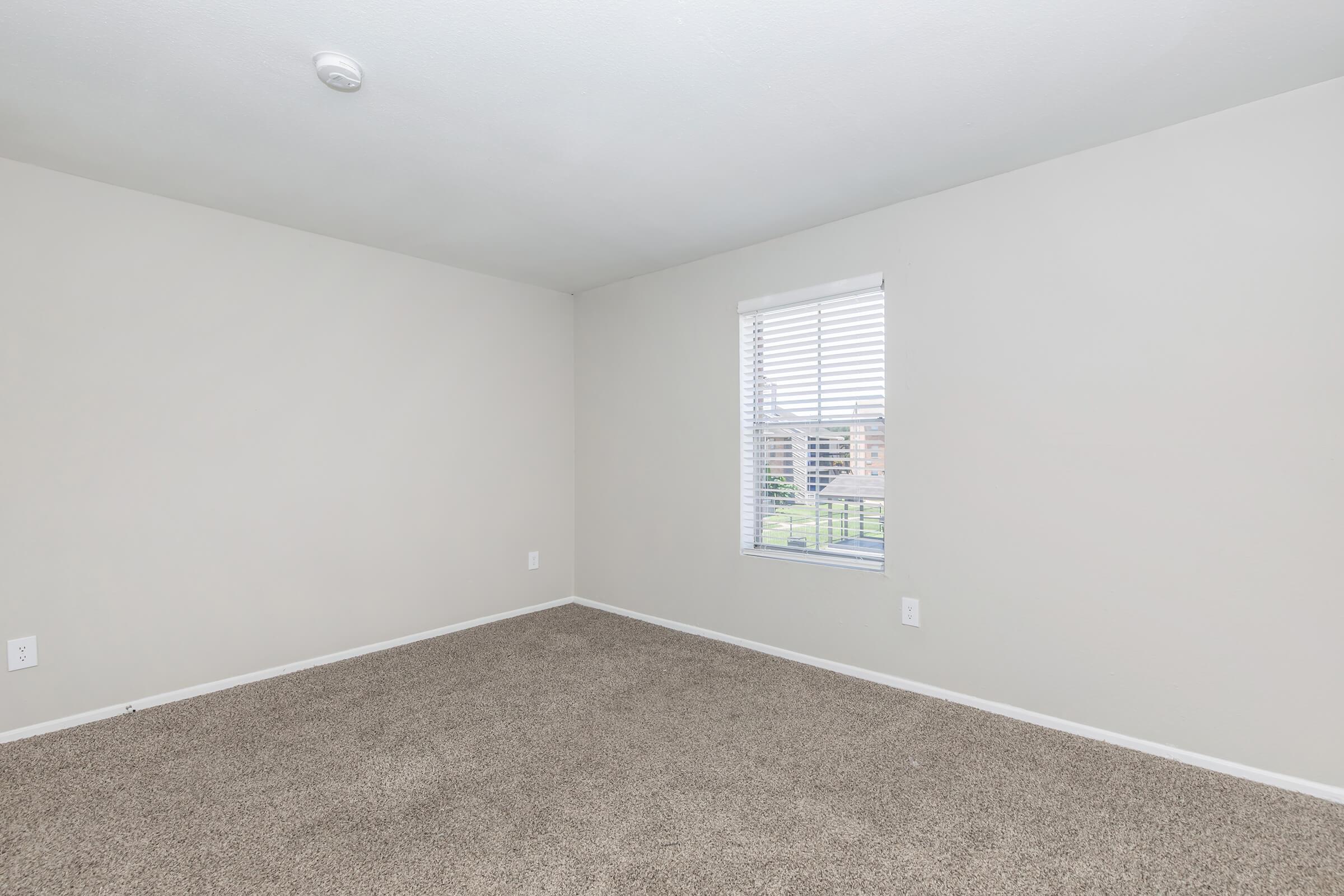 A vacant room with light beige walls and a window covered by white blinds. The floor is carpeted in a similar beige shade. There are no furnishings or decorations, creating a spacious and minimalistic atmosphere. A small ceiling light is visible.