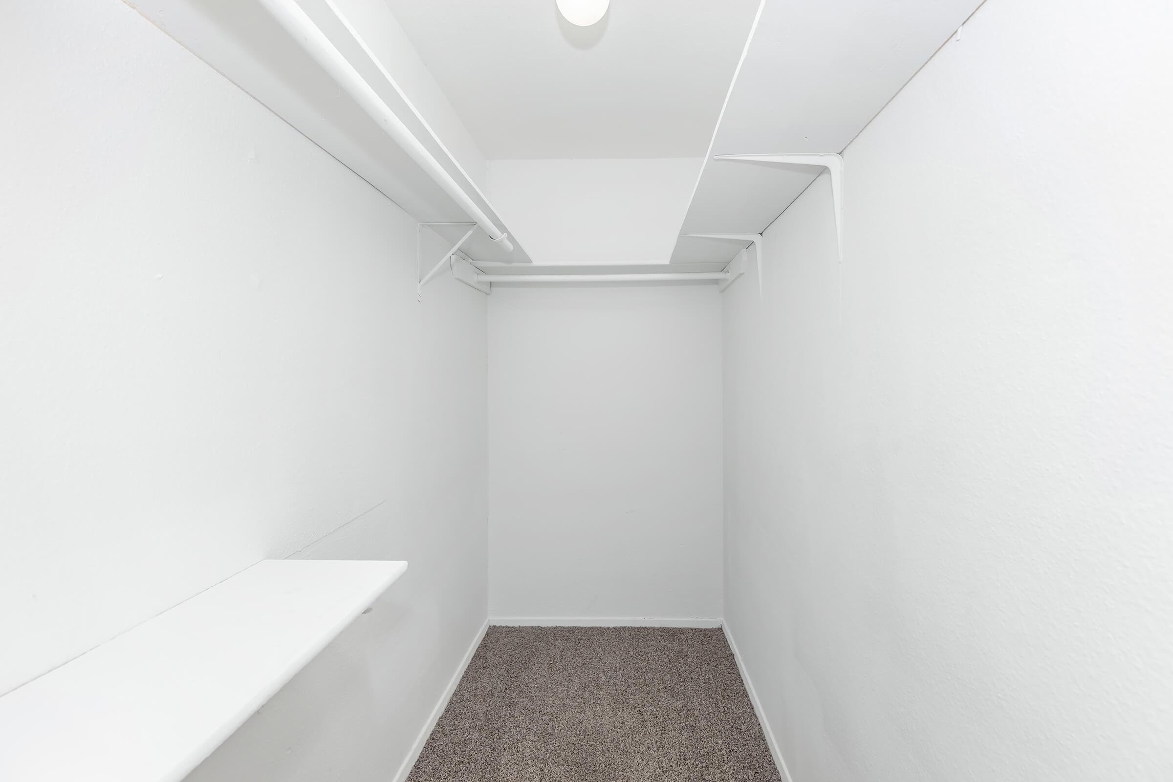 A small, empty closet with white walls and a light-colored carpet. There are two shelves along the left side and overhead, providing storage space. The room appears bright and minimally decorated, emphasizing its open and spacious feel despite its compact size.