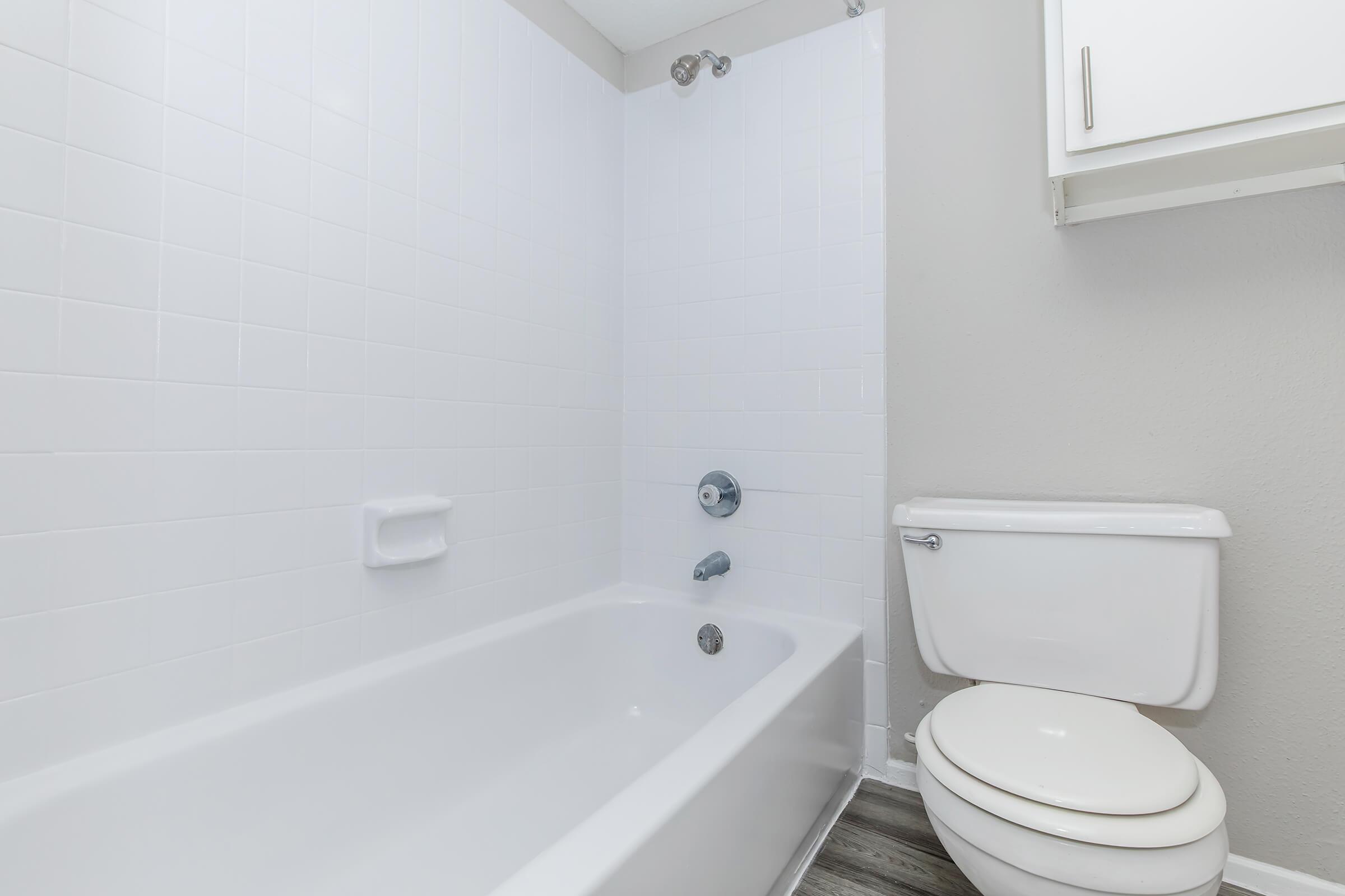 A clean, modern bathroom featuring a white bathtub with a shower, a flush toilet, and light gray walls. The space is well-lit, with a simple design and tile flooring, creating a fresh, minimalist appearance.