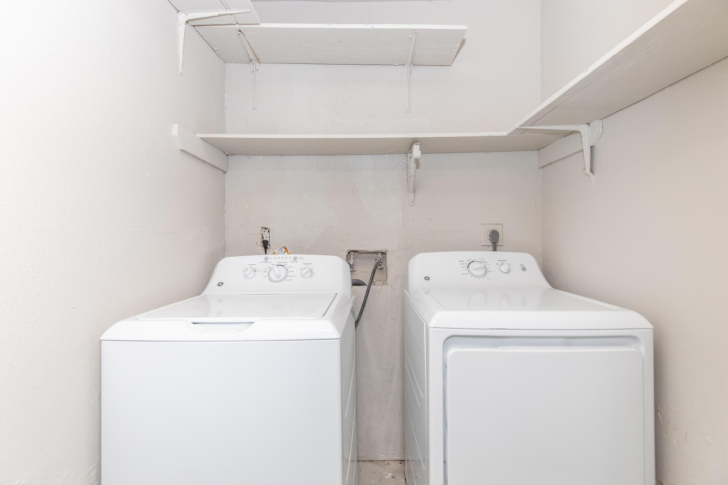 A clean laundry room featuring two white washing machines side by side, with simple shelving above them. The walls are painted white, creating a bright and tidy environment for laundry tasks.