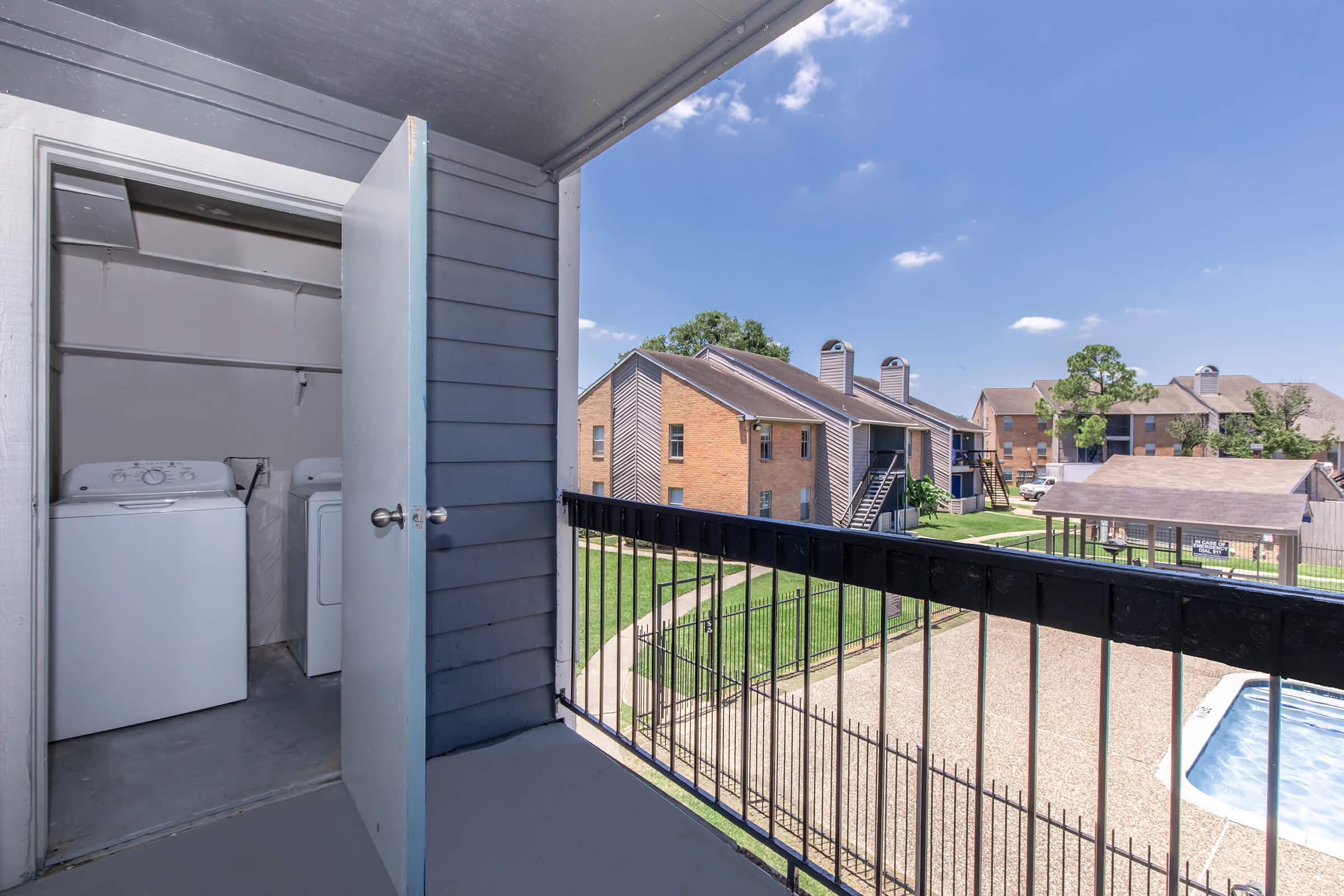 View from a balcony showing a laundry area with a washing machine and dryer, overlooking a courtyard with grassy spaces and apartment buildings. In the distance, there is a pool area bordered by a fence, under a clear blue sky.