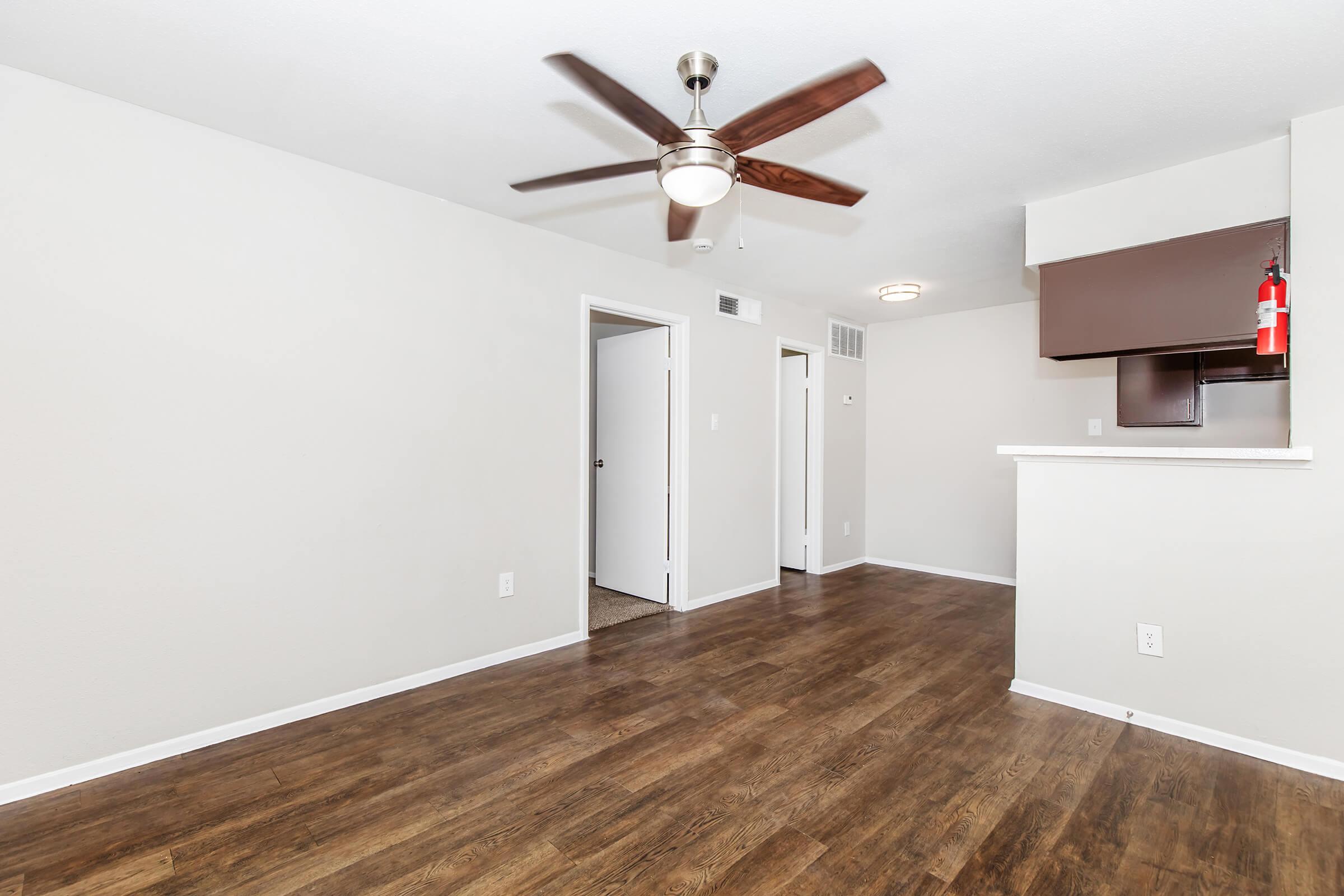 A spacious interior of an apartment featuring a wooden floor, a ceiling fan, and neutral-colored walls. There are two doorways leading to other rooms, and a partial view of a kitchen area with a bar counter. Natural light brightens the space, creating a welcoming atmosphere.