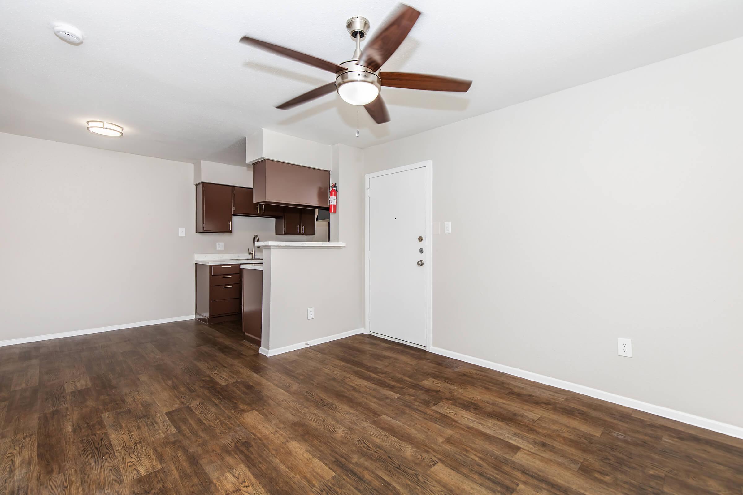 Spacious apartment interior featuring a living area with hardwood floors, a ceiling fan, and a white door leading outside. The kitchen area includes dark cabinetry and a countertop. Walls are painted light gray, creating a modern and inviting atmosphere.