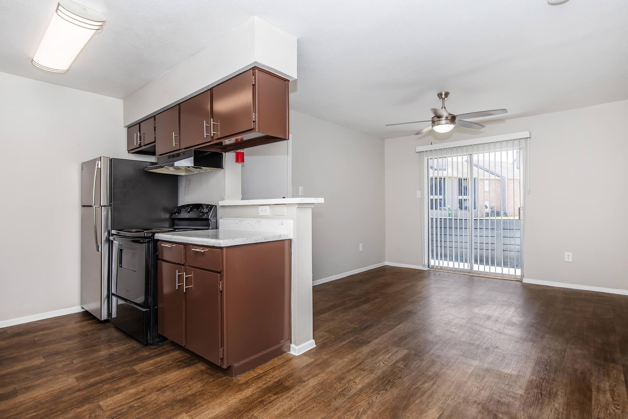 Modern kitchen with dark wood cabinets and stainless steel appliances, including a refrigerator and stove. Open layout leading to a living area with a ceiling fan and a sliding glass door that provides natural light and access to a balcony. Neutral-colored walls and wood-patterned flooring throughout.