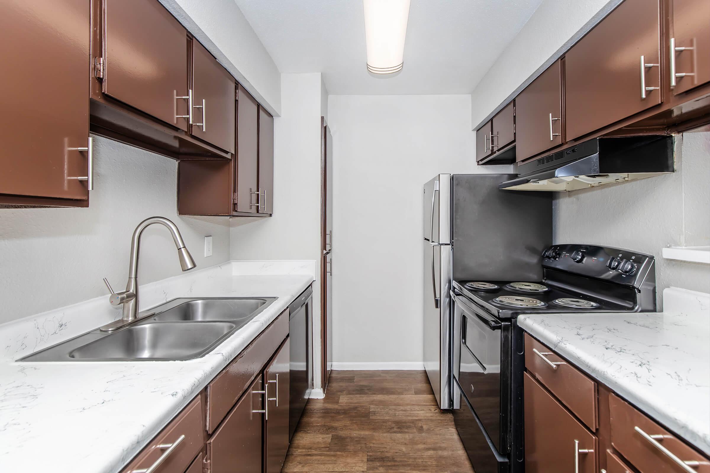 A modern kitchen featuring dark brown cabinets, a double sink, stainless steel appliances including a refrigerator and an oven, and a marble countertop. The floor is a dark wood laminate, and the walls are painted light gray, creating a bright and clean atmosphere.
