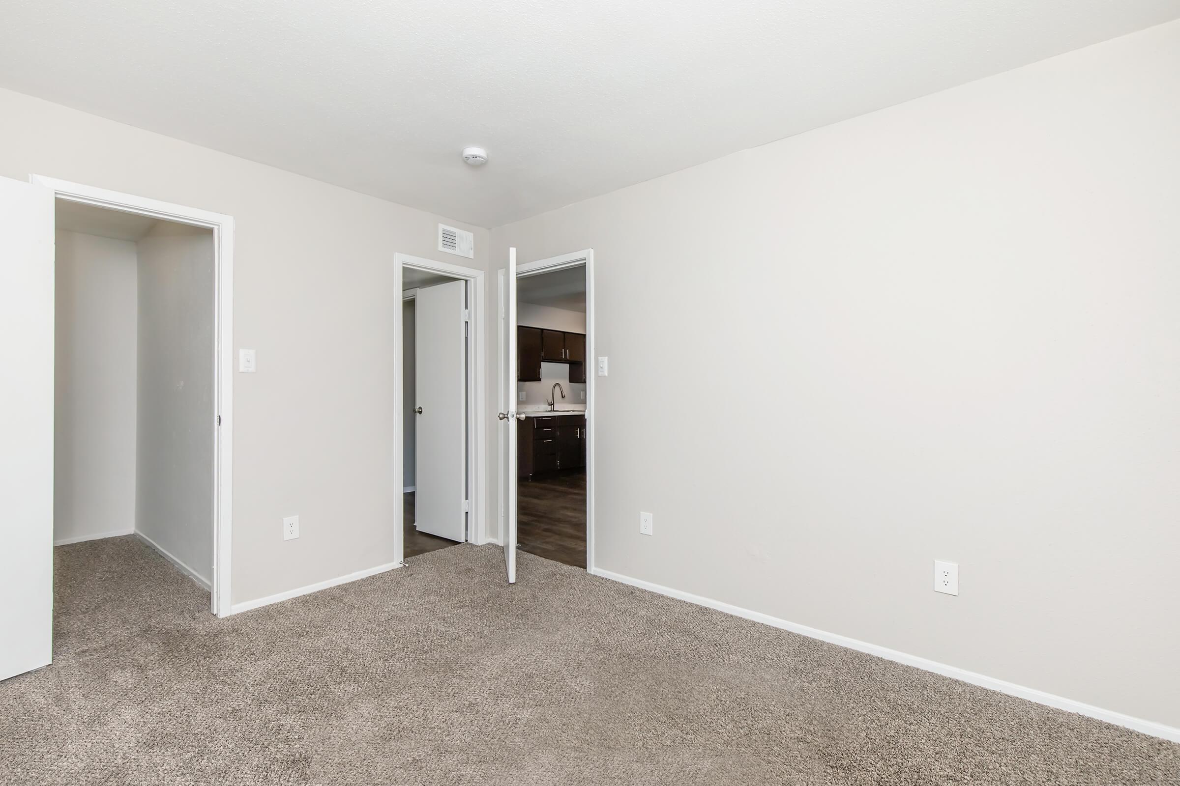 A view of an empty room with beige walls and carpeted flooring. There are two open doors leading to adjacent spaces, one appearing to lead to a small closet. The background features a glimpse of a kitchen area with dark cabinets, but the kitchen itself is not fully visible.