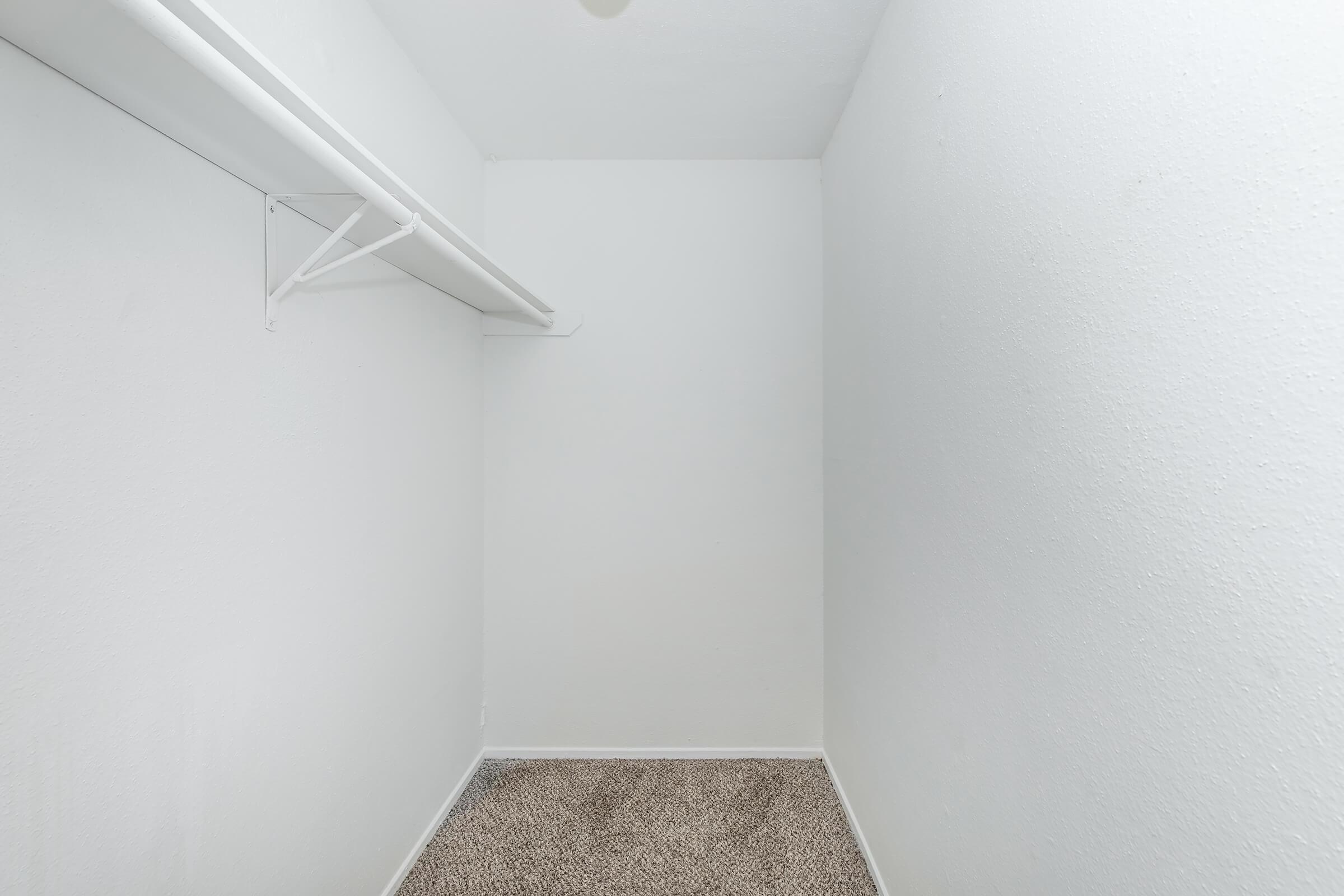Empty closet with white walls and a carpeted floor. There is a mounted shelf on the left side, and the space is well-lit, creating a clean and minimalistic appearance.