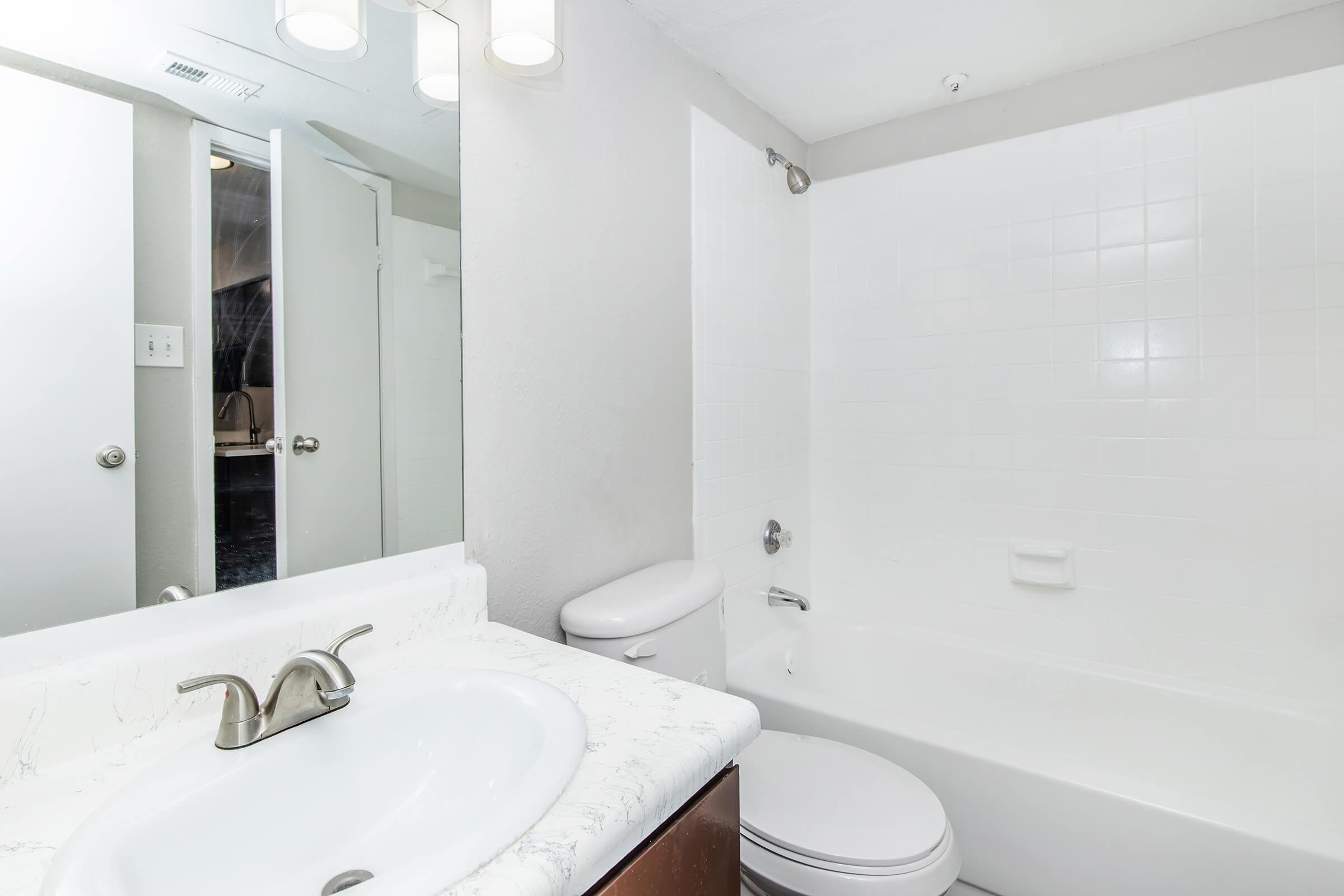 A clean, modern bathroom featuring a white tiled bathtub and shower, a sink with a marble countertop, and a large mirror. The bathroom includes a toilet and a door leading to another room. Bright lighting enhances the spacious feel of the area.