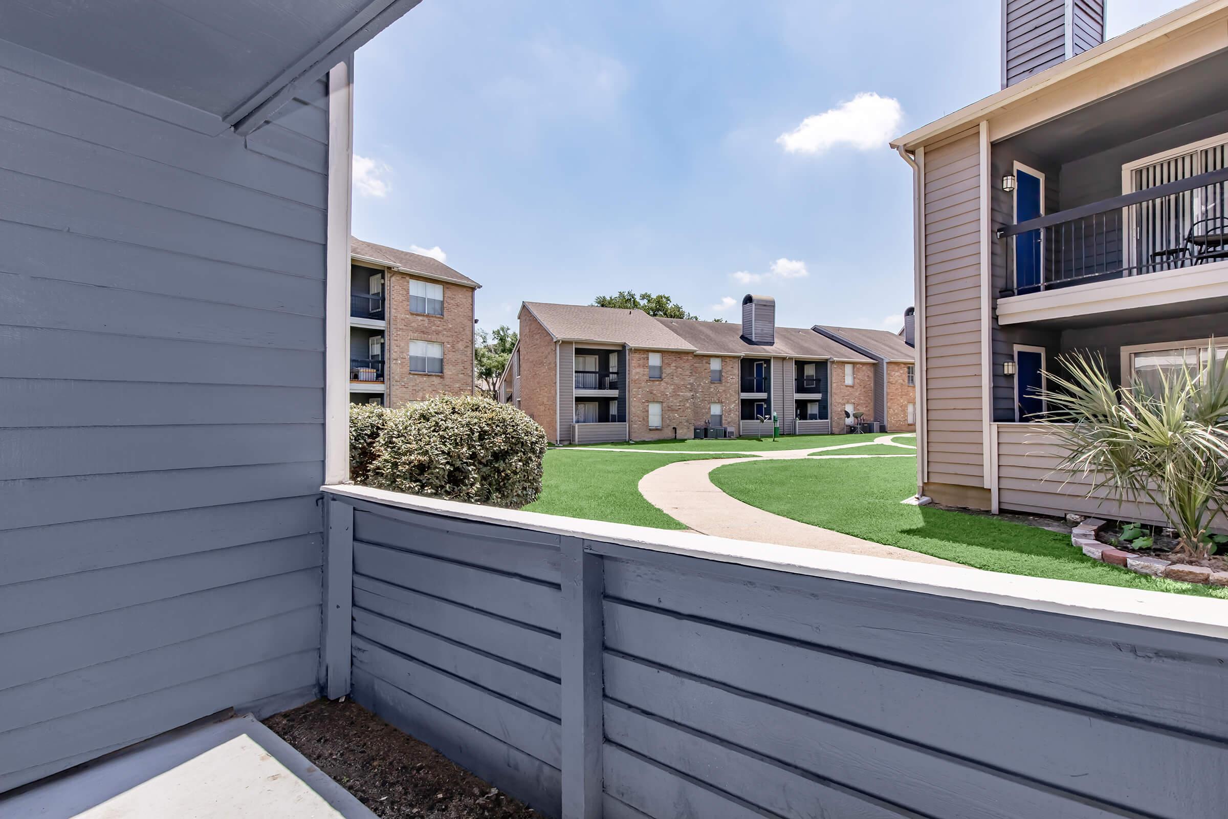 View from a balcony or porch showing a green lawn and walking path surrounded by multi-story apartment buildings. The sky is clear with a few clouds, creating a bright and inviting outdoor scene.