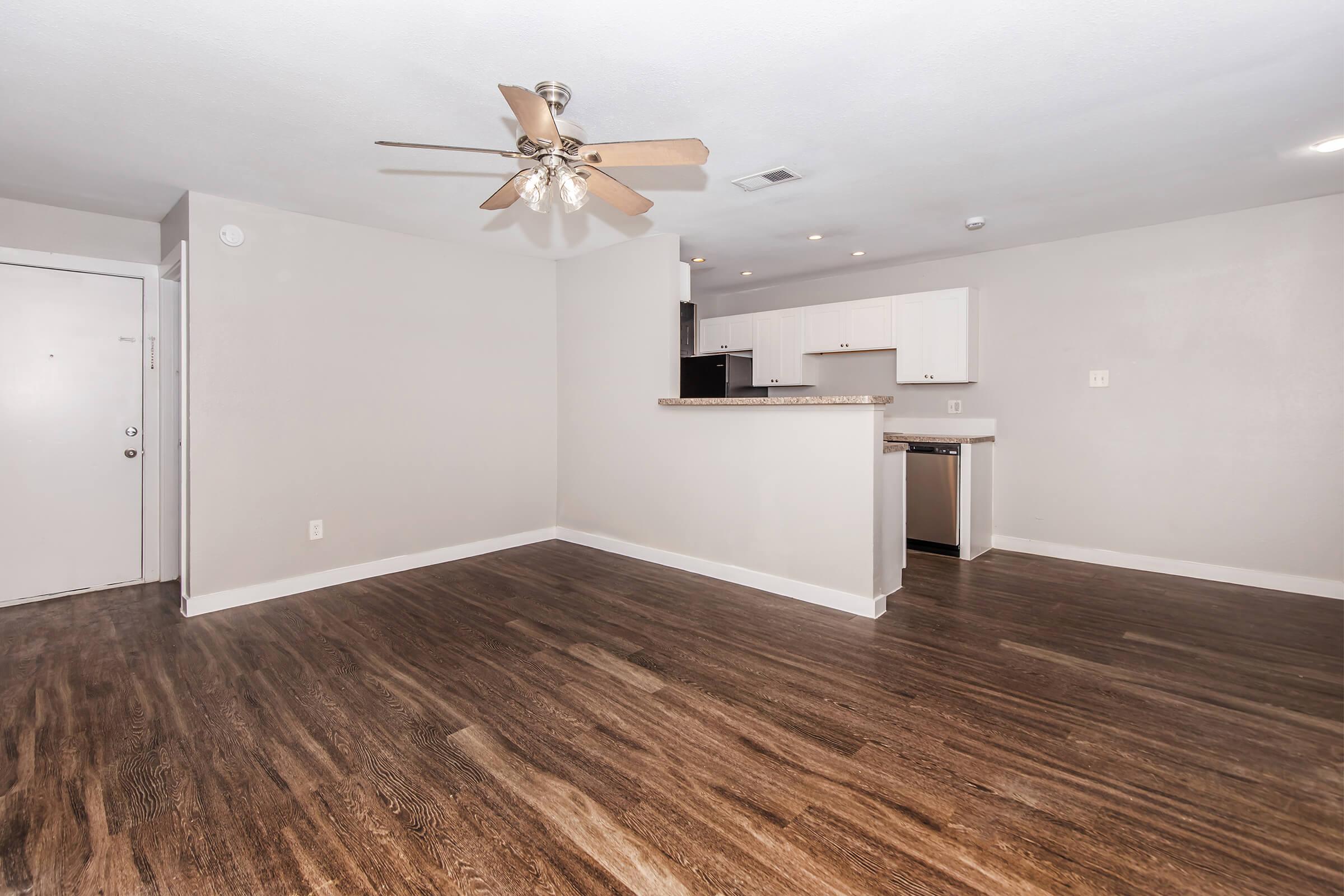 Interior view of a modern living space featuring a ceiling fan, neutral-colored walls, and hardwood-style flooring. The open layout includes a kitchen area with white cabinetry and stainless steel appliances, and a front door visible in the background. Natural light fills the room, creating a bright atmosphere.
