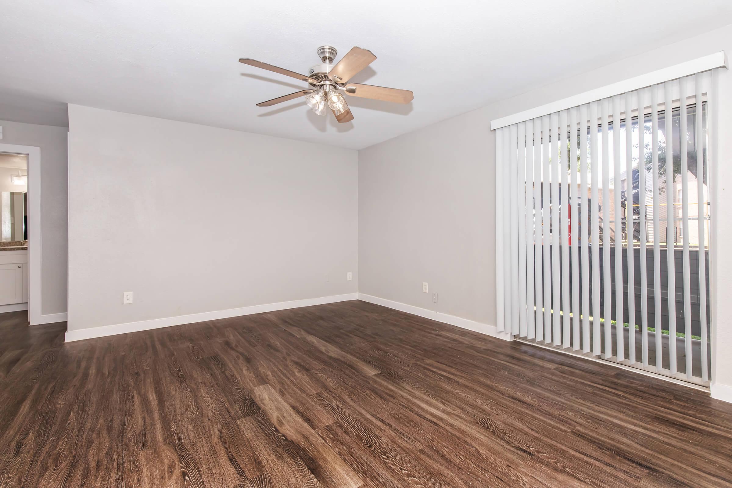A spacious, empty living room with light brown laminate flooring, a ceiling fan with wooden blades, and a large window with vertical blinds allowing natural light. A doorway on the left leads to another room, and the walls are painted a light gray, creating a bright and airy atmosphere.