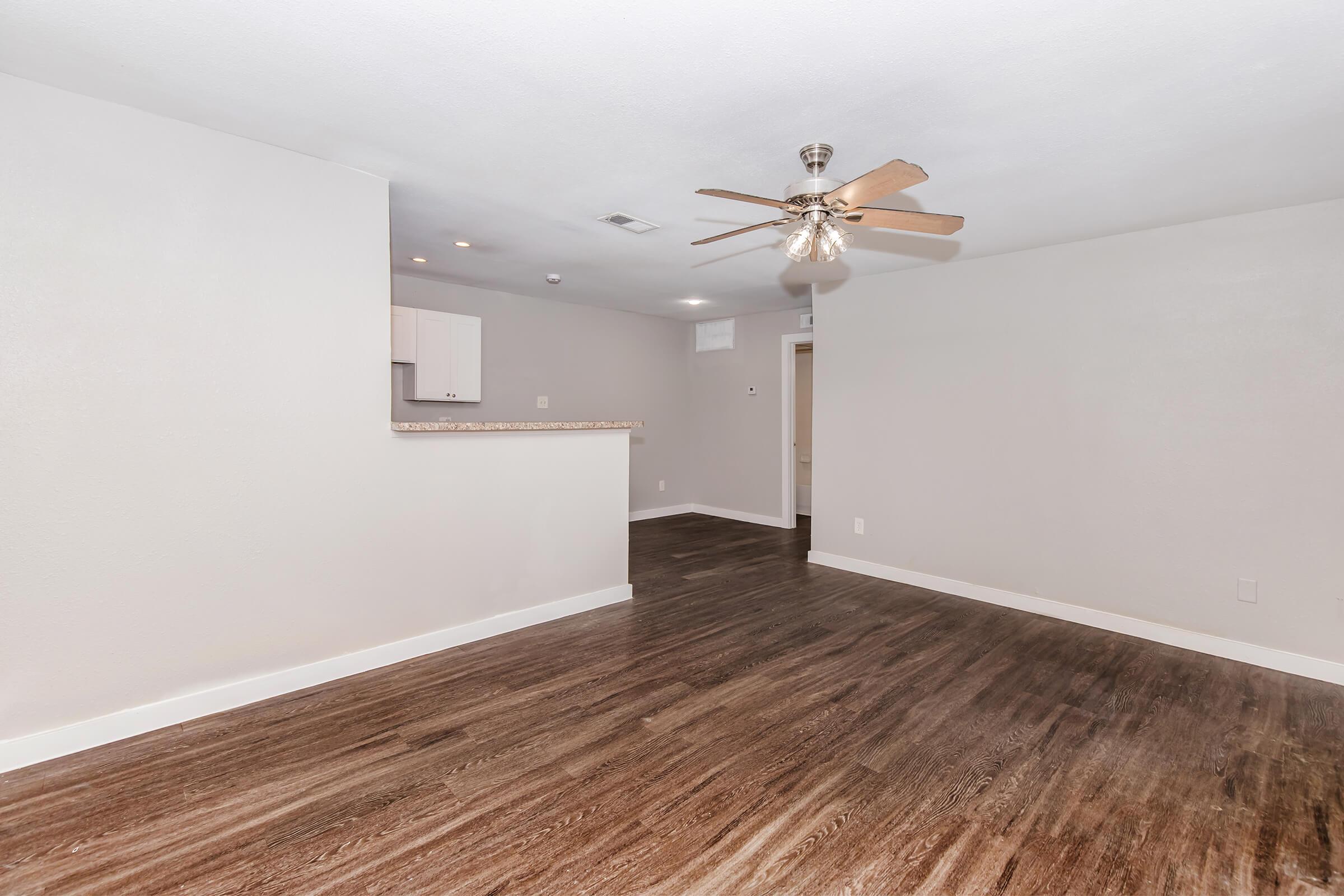A spacious, modern living area with light-colored walls and dark hardwood flooring. The room features a ceiling fan and an open layout, leading to a kitchen area with a breakfast bar. Doorways to other rooms are visible, creating a bright and inviting atmosphere.