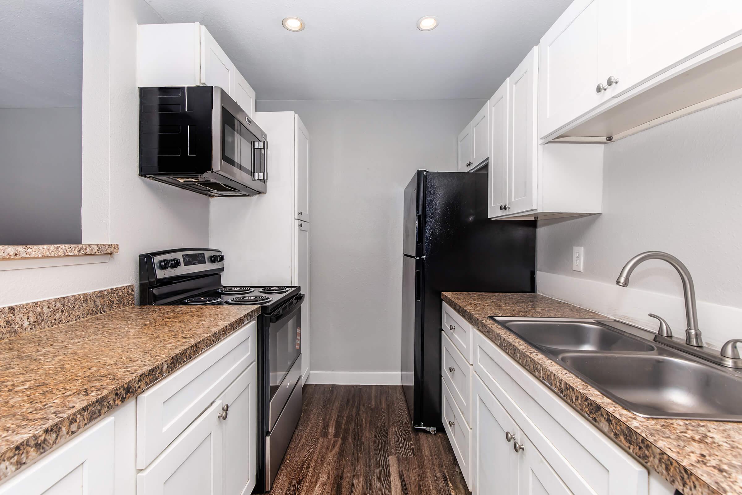 A modern kitchen featuring white cabinetry, granite countertops, and stainless steel appliances, including a stove, microwave, and a refrigerator. The kitchen has a sleek design with an open layout, good lighting, and hardwood-style flooring.