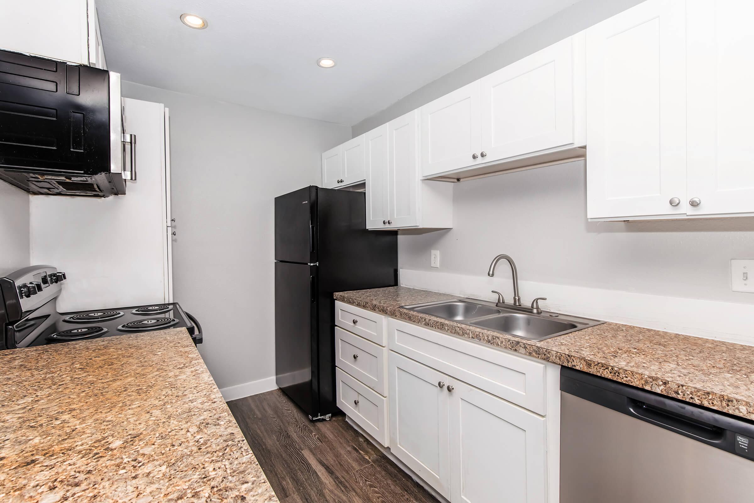 Modern kitchen with white cabinetry, granite countertops, and stainless steel appliances. Features a black refrigerator, a gas stove, dual sink, and a dishwasher. The walls are painted light gray, and the flooring is dark wood laminate. Well-lit with recessed ceiling lights.