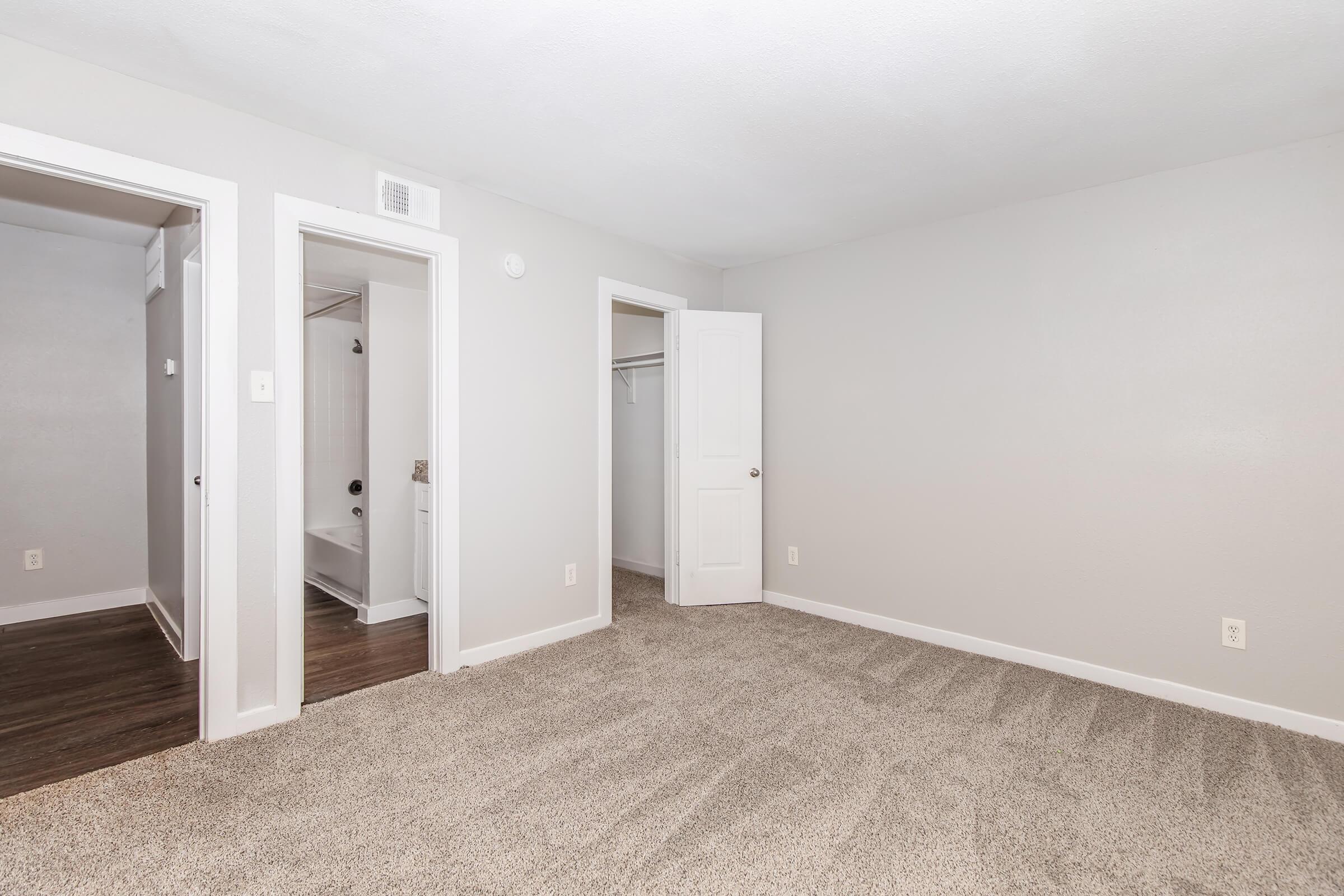 A spacious room with freshly painted gray walls and plush carpeting. The room features two doorways leading to adjacent spaces, one with a closet and the other with a bathroom. Natural light enters the room, creating a bright and inviting atmosphere.
