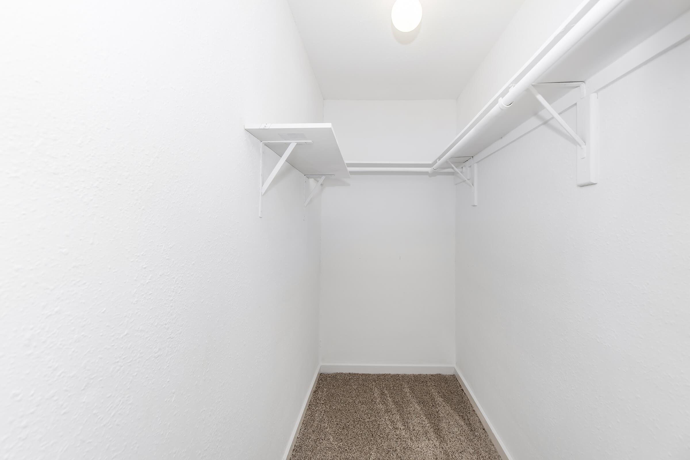 Empty closet with white walls and a single overhead light. There is a shelving unit on the left side supported by brackets, and the floor is covered with beige carpeting. The space appears clean and unoccupied, providing a minimalistic storage area.