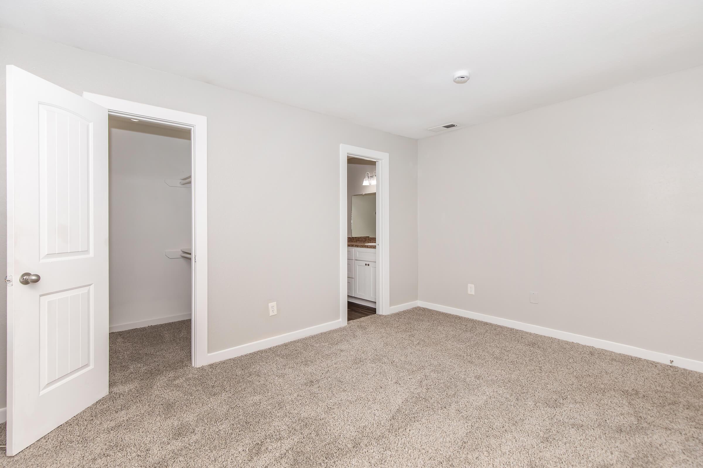 A spacious, empty room with beige carpet and light gray walls. There are two doors: one leading to a hallway and the other to an adjacent bathroom. The lighting is bright, highlighting the simplicity of the space, which is suitable for various uses like a bedroom or office.