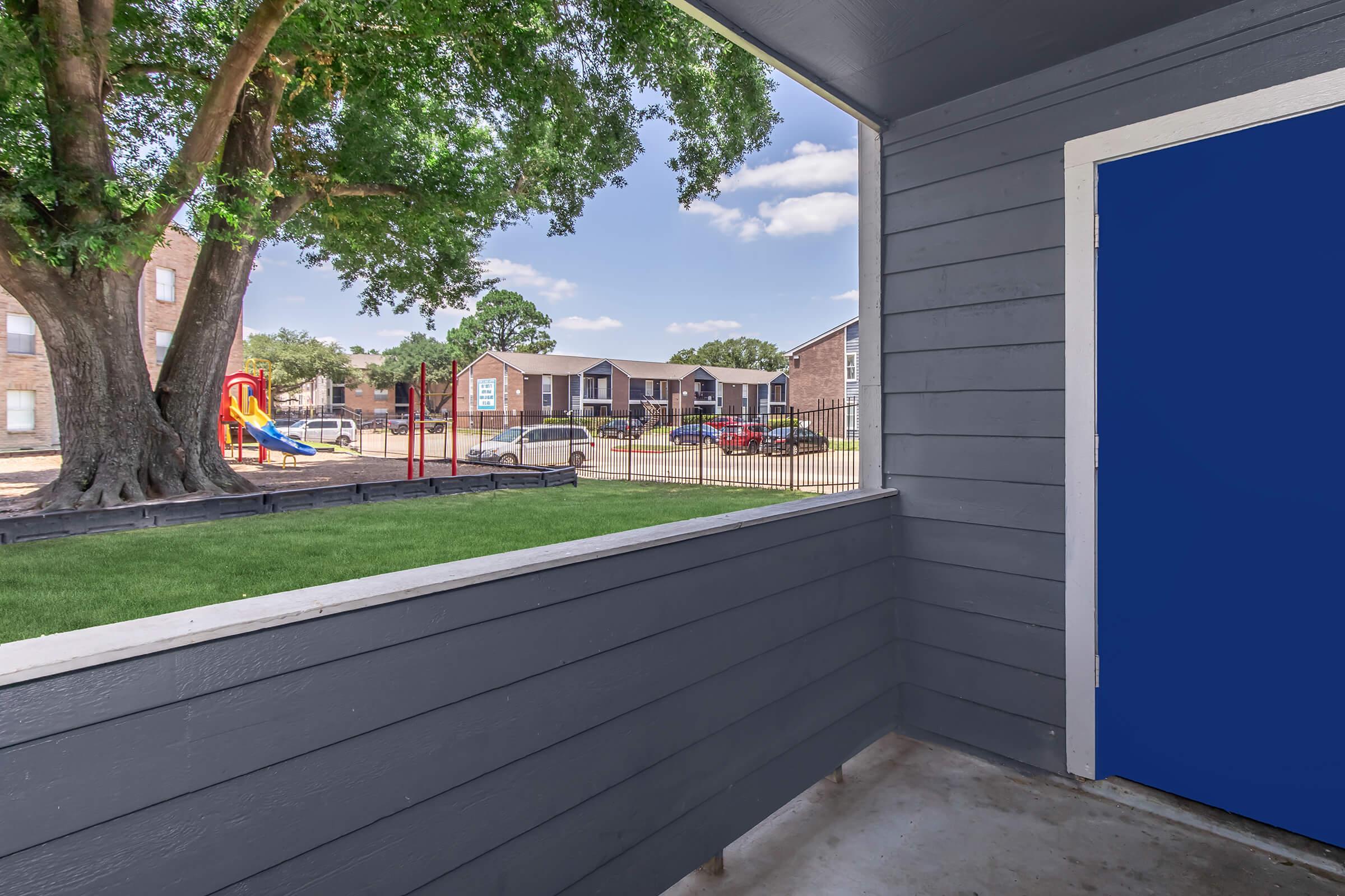 View from a covered porch showing a playground and apartment buildings in the background. Bright green grass is visible in the foreground, along with a blue door on the right side of the image. The sky is partly cloudy.