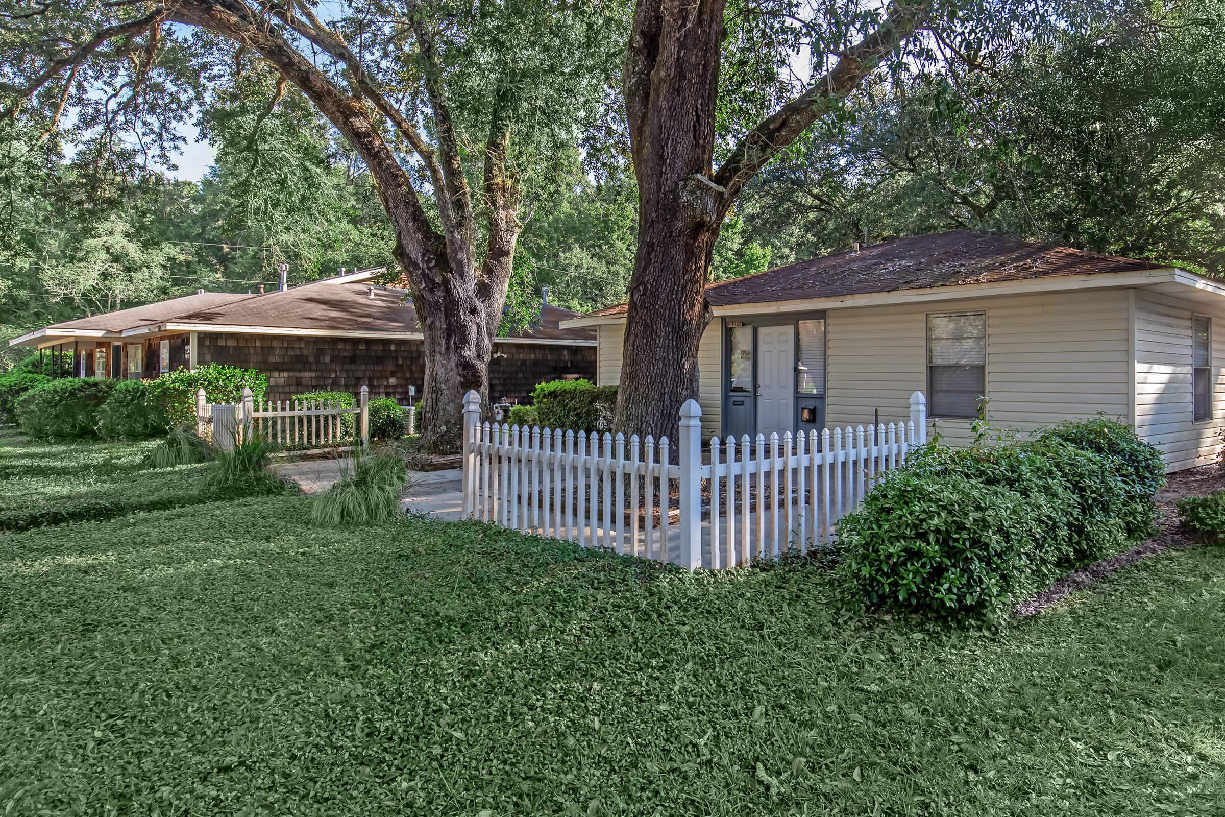 A serene neighborhood scene featuring two homes with natural surroundings. The houses are lined with green lawns and white picket fences, shaded by large trees. The atmosphere is calm and inviting, suggesting a peaceful residential area.