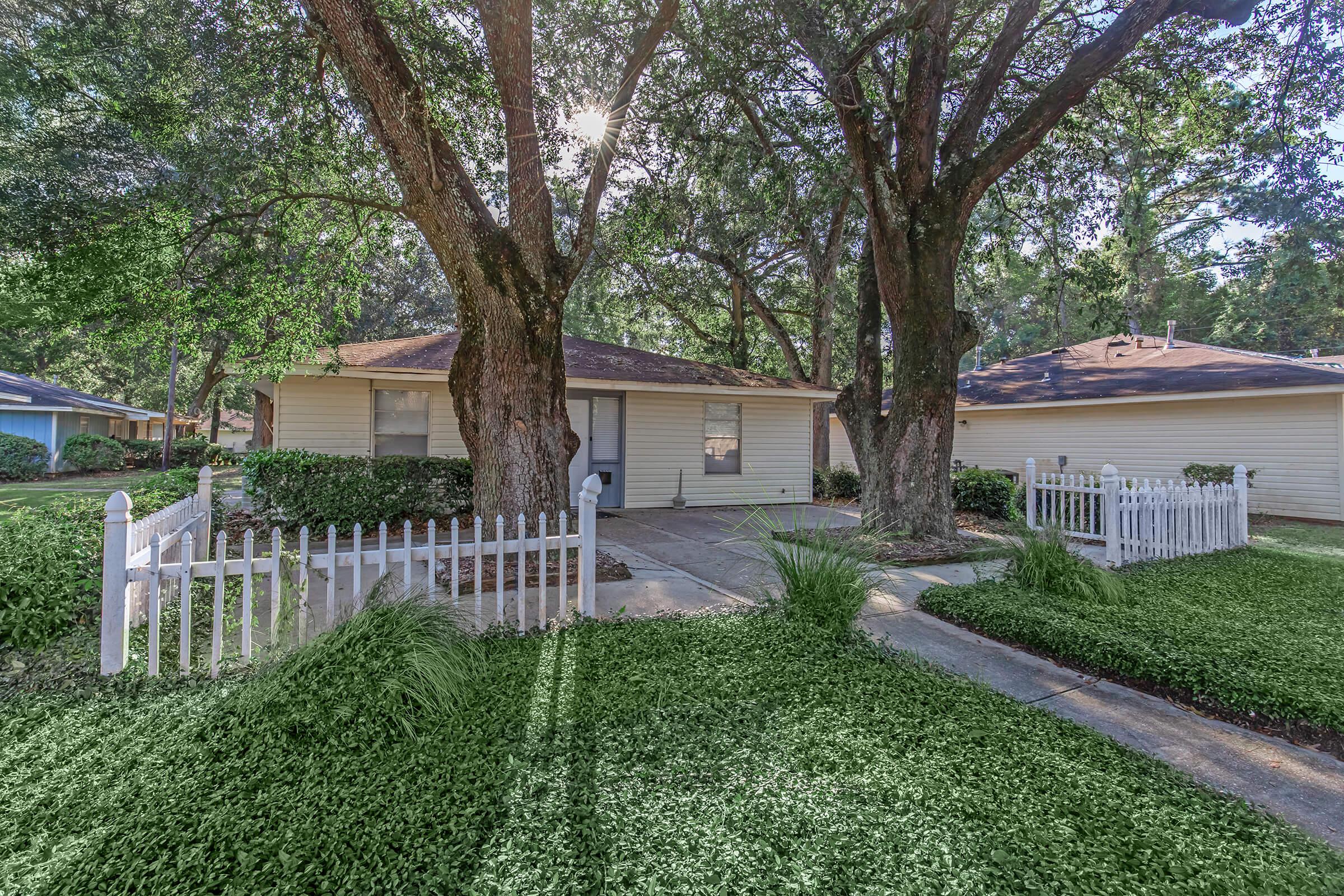 A quaint house surrounded by lush green grass and two large trees, with a white picket fence enclosing the front yard. Sunlight filters through the leaves, creating a warm and inviting atmosphere. The driveway leads to the entrance, with nearby homes visible in the background.