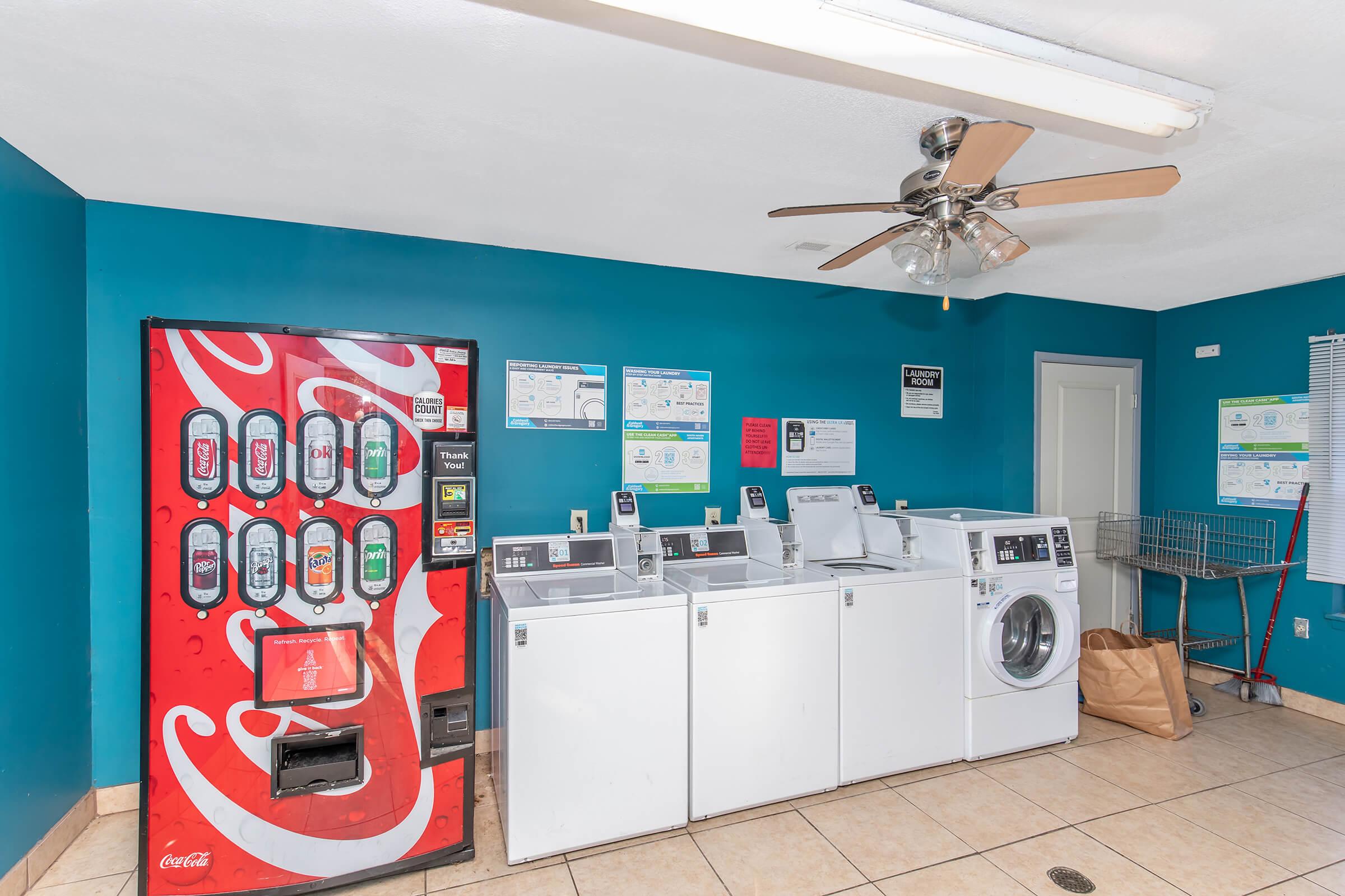 A laundry room featuring a Coca-Cola vending machine on the left. There are several washing machines and a dryer in the middle, with a ceiling fan above. The walls are painted blue, and there are various posters and signs related to laundry services displayed on the wall. A laundry basket can be seen in the corner.
