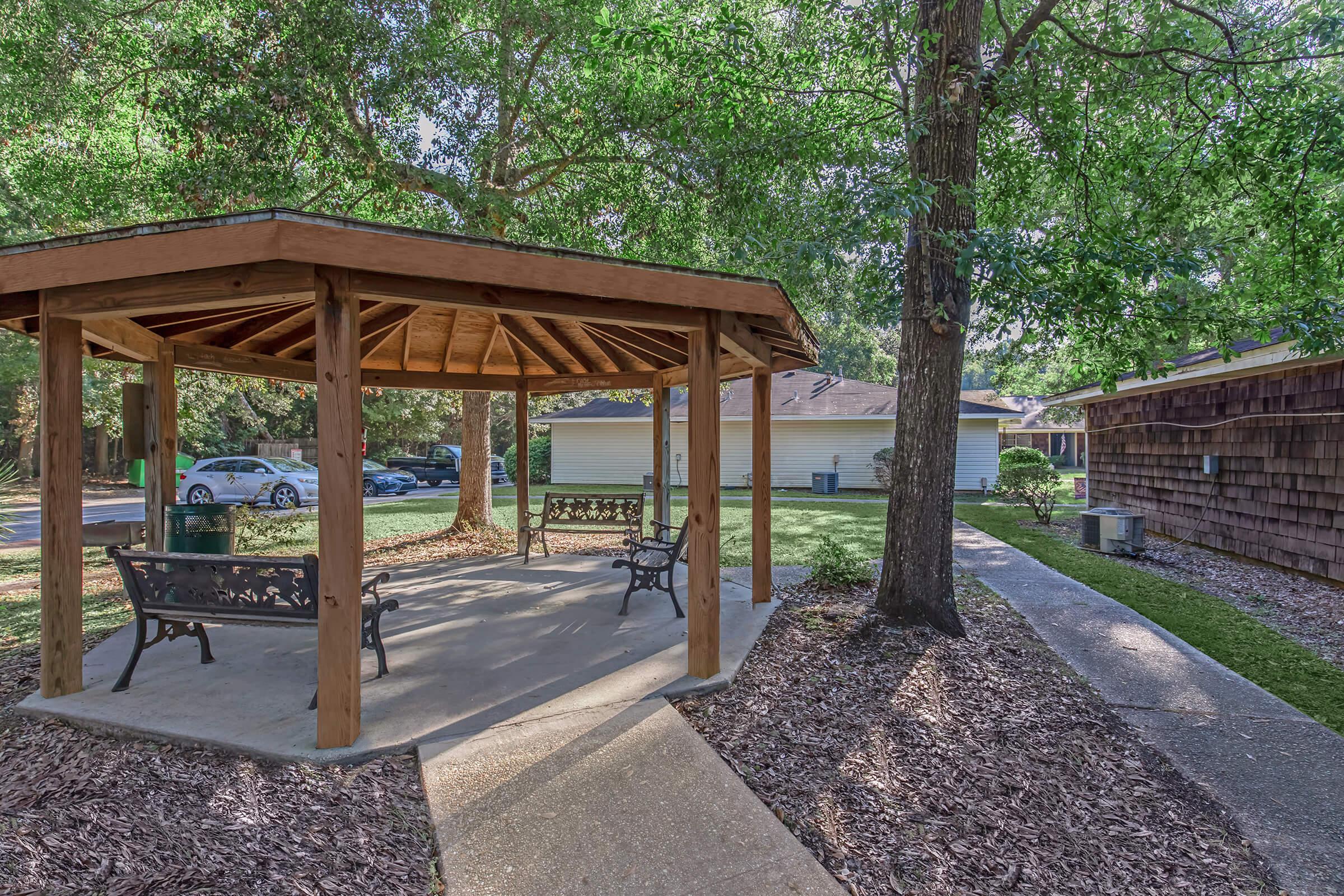 A wooden gazebo with benches, surrounded by lush green trees and mulch. Nearby, there are two houses and parked cars, with a concrete pathway leading through the area. The setting is peaceful and well-maintained, providing a relaxing outdoor space.
