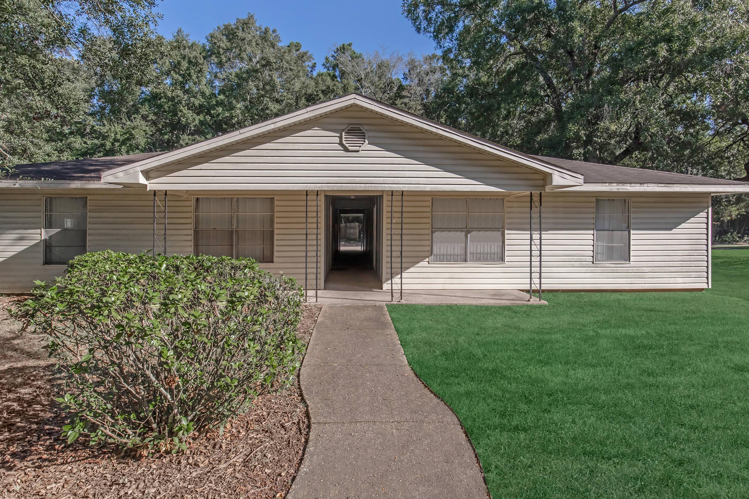 A single-story house with a light-colored exterior and a simple roof. The front yard features a neatly trimmed green lawn and a pathway leading to the entrance. Trees are visible in the background, providing shade and greenery. The house has several windows and is set in a spacious, well-maintained environment.